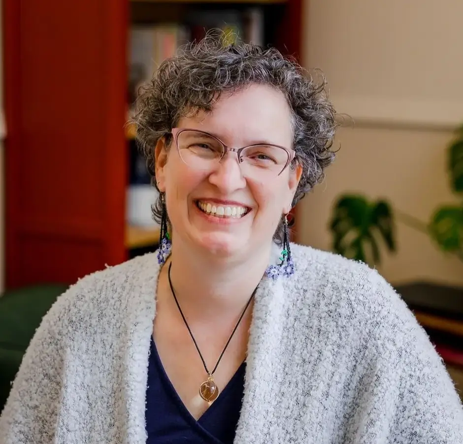 Dr Amy Kelchner, with short curly gray hair, glasses, and colorful earrings, smiles warmly. She wears a gray textured cardigan, a black top, and a heart-shaped pendant. A plant and bookshelf are visible in the softly lit background.
