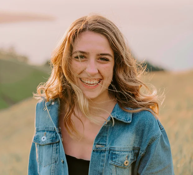 A young woman with wavy light brown hair smiles outdoors, wearing a blue denim jacket. Soft sunlight and a blurred natural landscape set a serene scene, evoking the calm often sought through ketamine therapy.