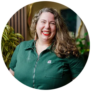 A woman with long wavy hair, wearing a dark green collared shirt, smiles broadly while standing indoors near plants—her positive energy reflecting the hope often found through ketamine therapy.