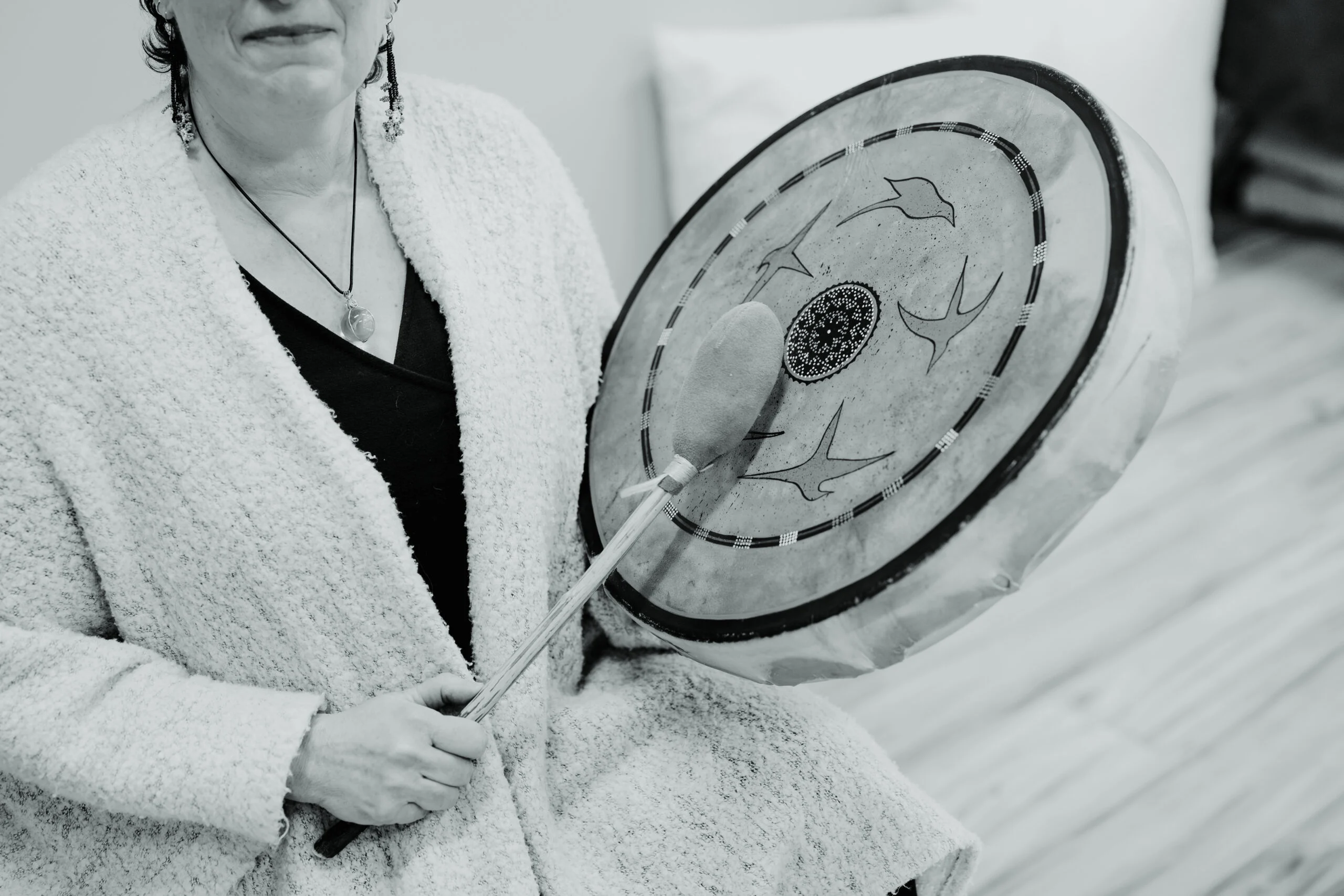 Dr Amy Kelchner, shown in this black and white photo, wears a textured sweater while holding a decorated hand drum and beater. The drum’s design features birds in flight, capturing her artistic spirit.