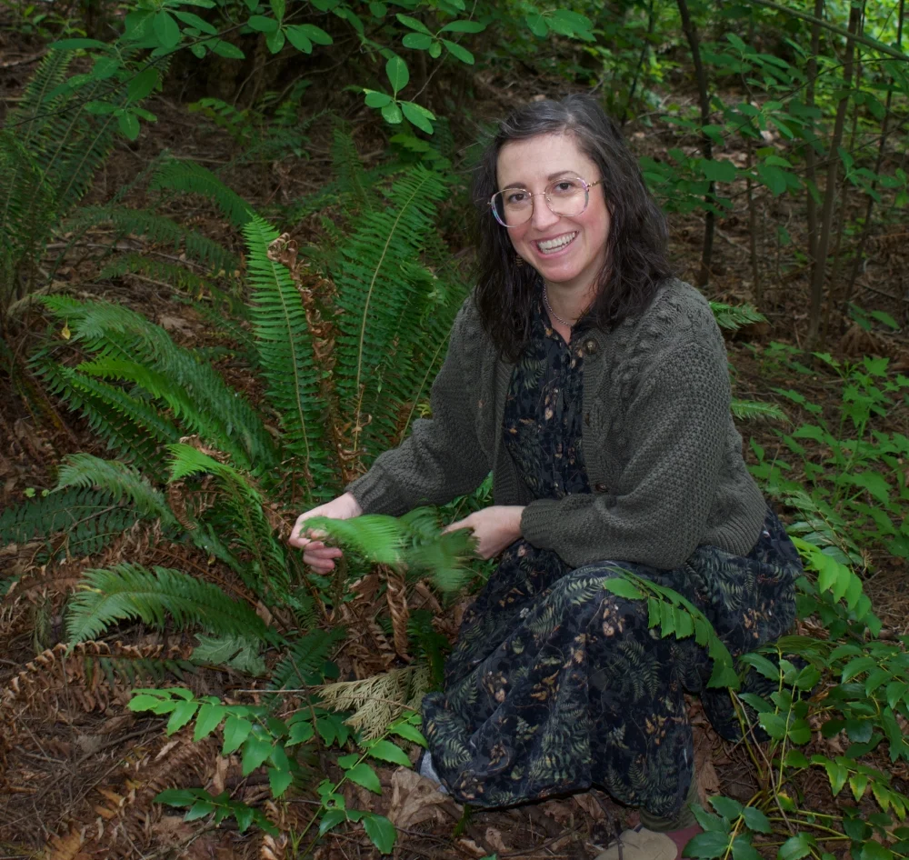 Dr Kara Crisp, a woman with glasses and dark hair, wearing a patterned dress and sweater, kneels in a forest, smiling and holding the fronds of a green fern surrounded by leafy plants and trees.