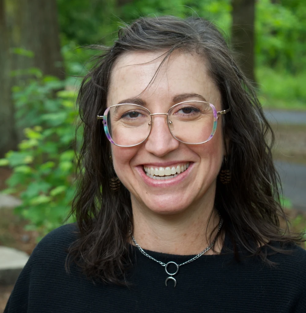 Dr Kara Crisp, a woman with wavy brown hair and glasses, smiles outdoors in a black top, crescent moon necklace, and brown earrings. Green foliage and tree trunks form the natural background.