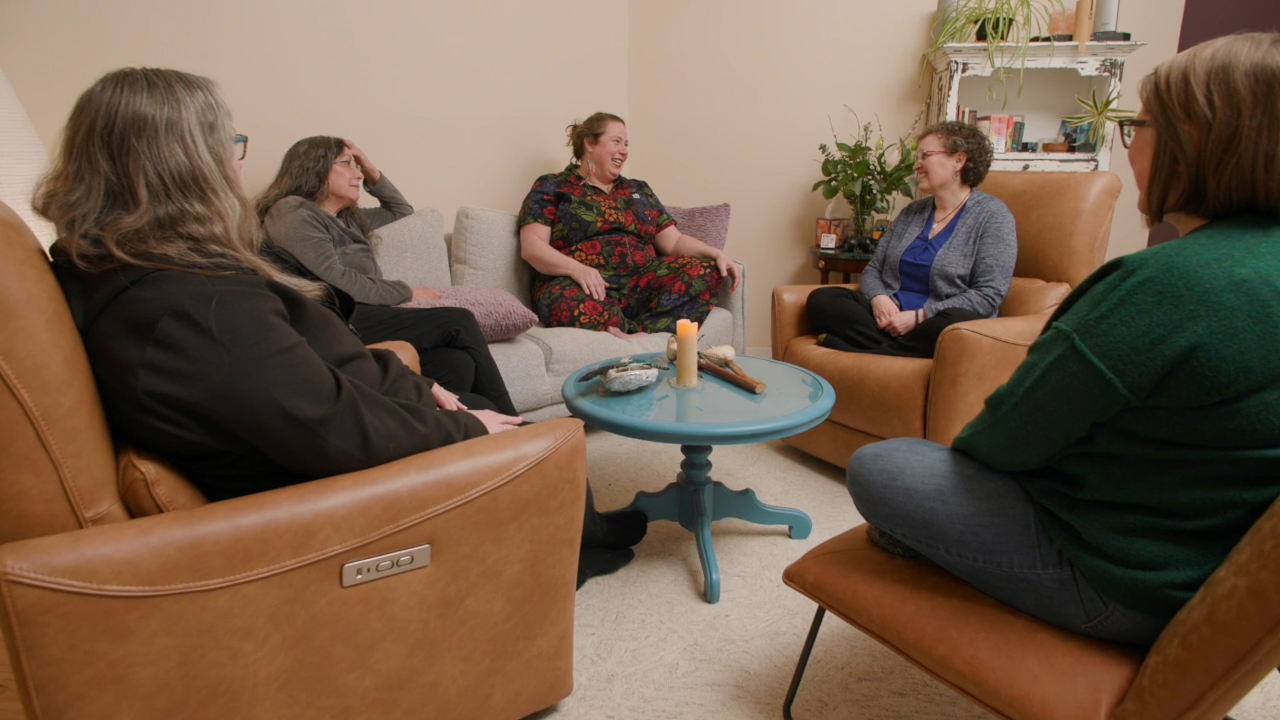 Five women sit in a cozy living room on chairs and a sofa, arranged in a circle around a small blue table with candles and decor. As they discuss ketamine therapy, they appear relaxed and cheerful, enjoying an open and supportive conversation.