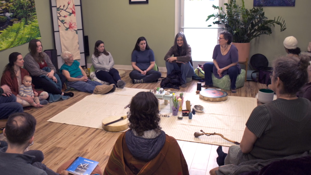 A group of people sits in a circle on the wooden floor of a green-walled room, surrounded by plants, drums, and ritual objects, engaged in a group discussion or ketamine therapy ceremony.