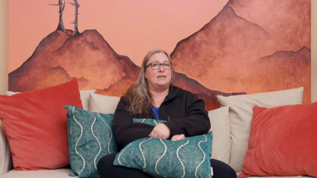 A woman with long hair and glasses sits on a beige couch with red and teal patterned pillows, reflecting on her ketamine therapy journey, in front of a mural featuring orange and brown mountains.