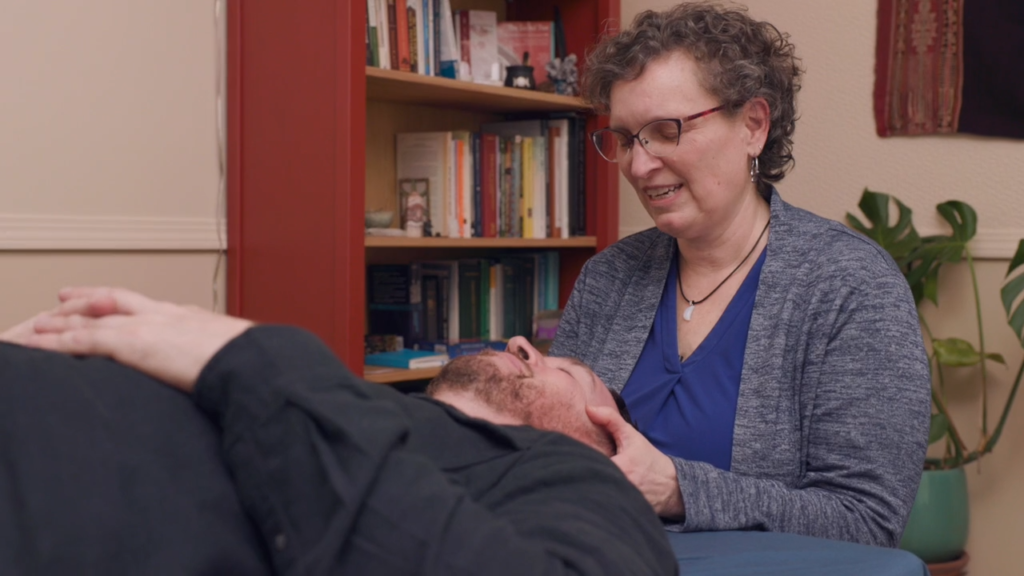 A woman with curly hair and glasses smiles while gently holding the head of a man lying down, in a room with bookshelves—suggesting a calming environment often used for ketamine therapy.