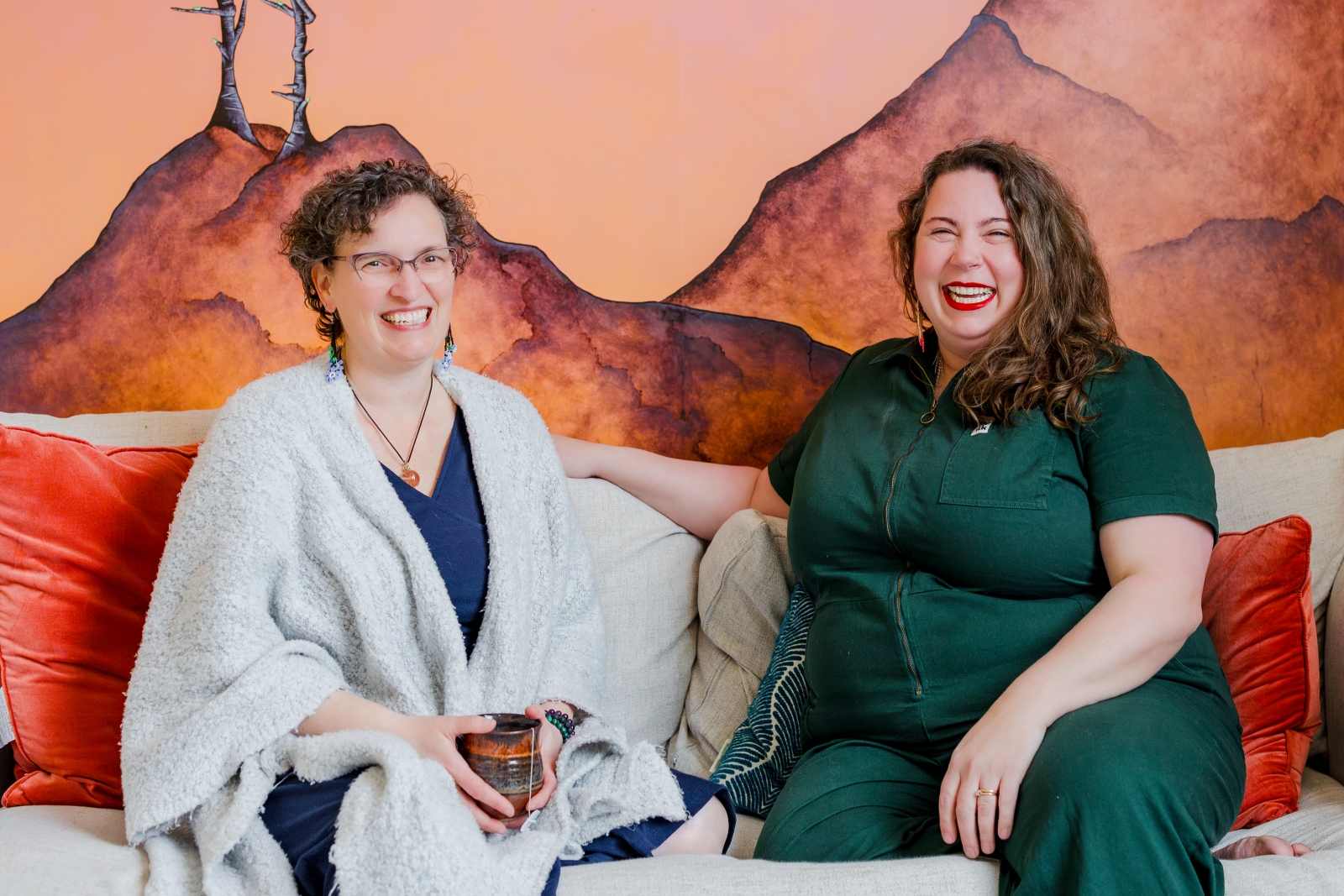 Two women sit together on a beige couch with orange cushions, smiling and laughing after a ketamine therapy session. One wears a green jumpsuit, the other a blue dress with a light shawl, holding a mug. A mural of mountains is painted behind them.