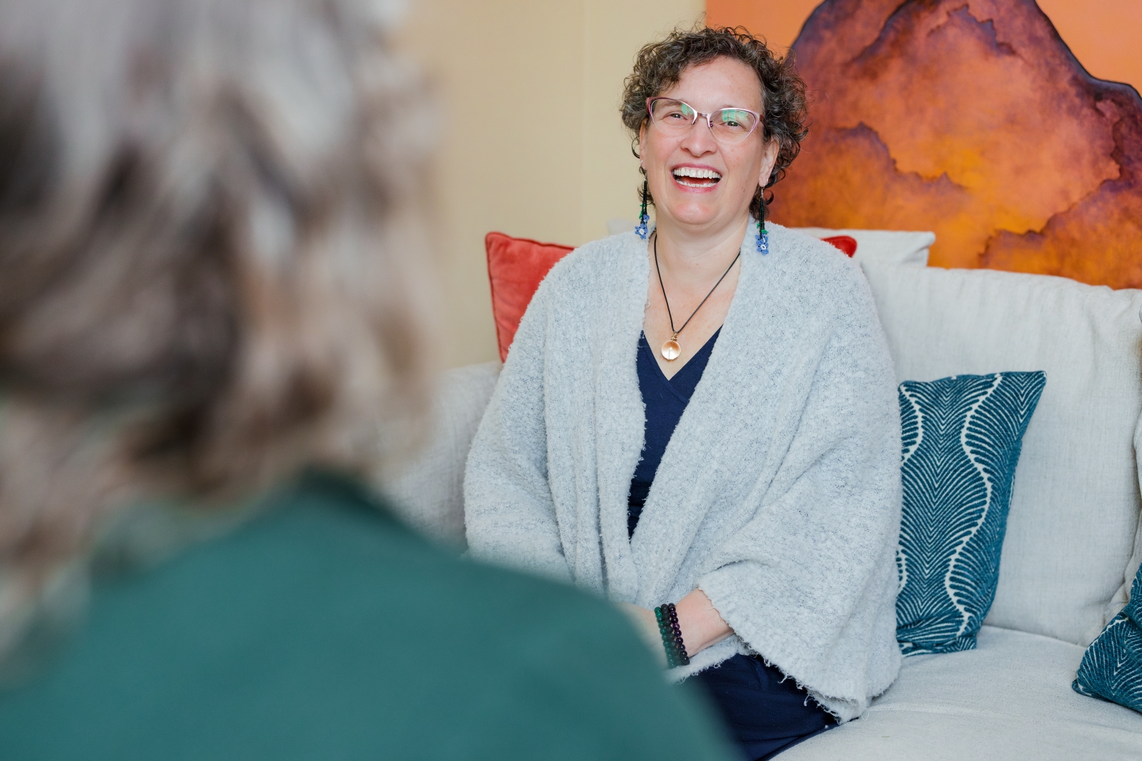 A woman with curly hair and glasses, wearing a light gray shawl, sits on a couch with patterned blue pillows, smiling and discussing ketamine therapy with another person whose back is to the camera.