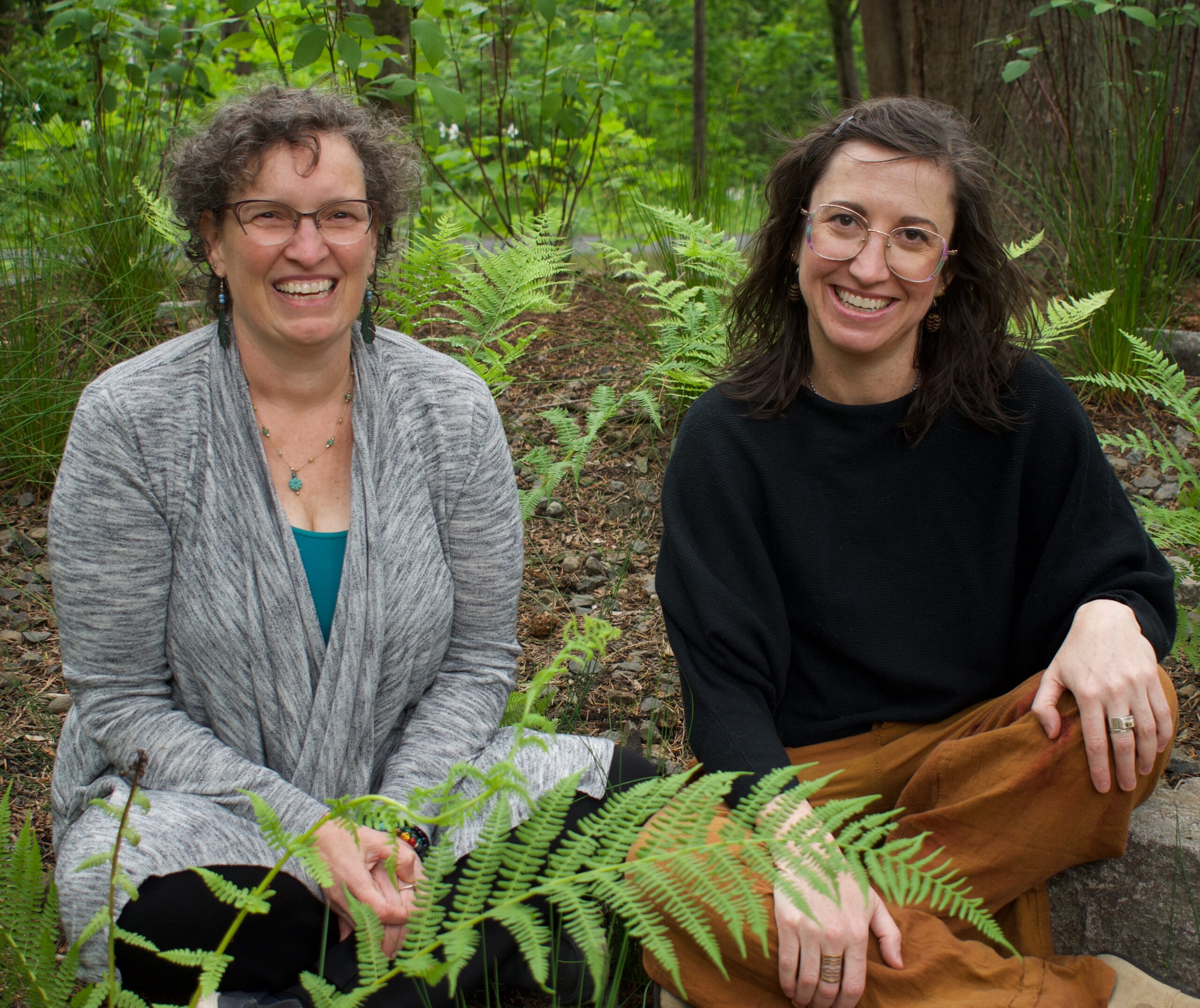Two women sit outdoors among green ferns and trees, smiling at the camera. They appear relaxed and content, reflecting the calm often sought through ketamine therapy. One wears glasses and a gray cardigan; the other wears a black sweater and brown pants.