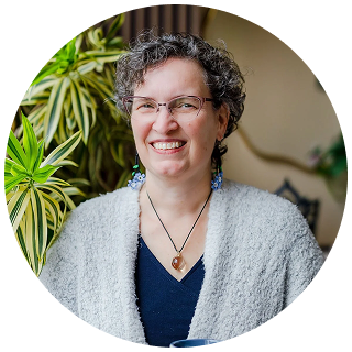 A smiling person with short curly hair, glasses, and a gray sweater sits indoors near green leafy plants, holding a cup and wearing a pendant necklace—perhaps reflecting after a recent ketamine therapy session.