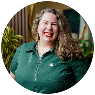 Dr Amy Kelchner, with long curly hair and a bright smile, wears a green button-up shirt while standing in an indoor setting filled with plants—perfect for a bio photo.