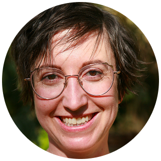 A person with short brown hair and glasses smiles at the camera in a close-up portrait, with a blurred outdoor background, embodying the calm confidence often seen after successful ketamine therapy.