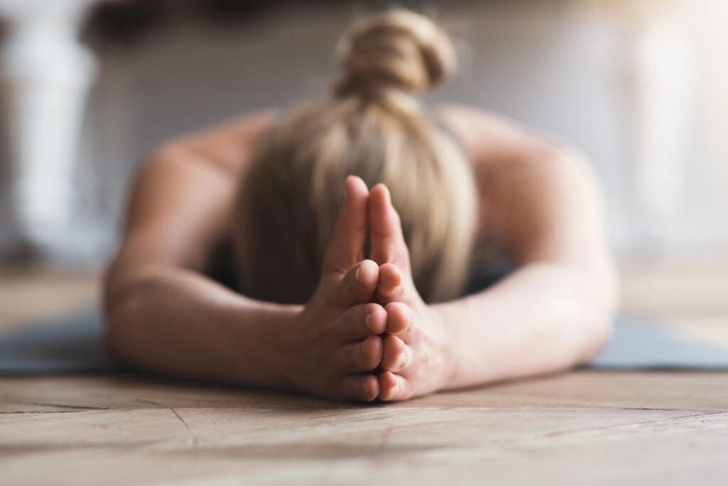 A person with blonde hair in a bun is stretching forward on a yoga mat, arms extended and palms pressed together, resting their forehead on the floor—capturing a tranquil moment that echoes the calm found in ketamine therapy.