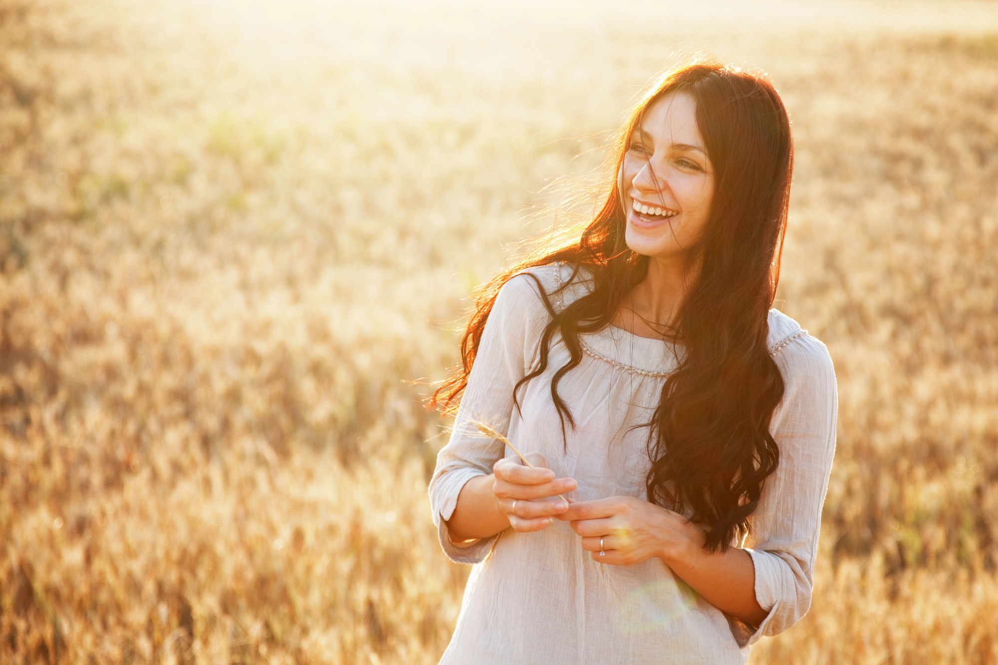 A young woman with long brown hair smiles while standing in a sunlit field of tall grass near Gresham, wearing a light-colored blouse. The background is warmly lit by the sun, evoking the calm often found after Ketamine Therapy.