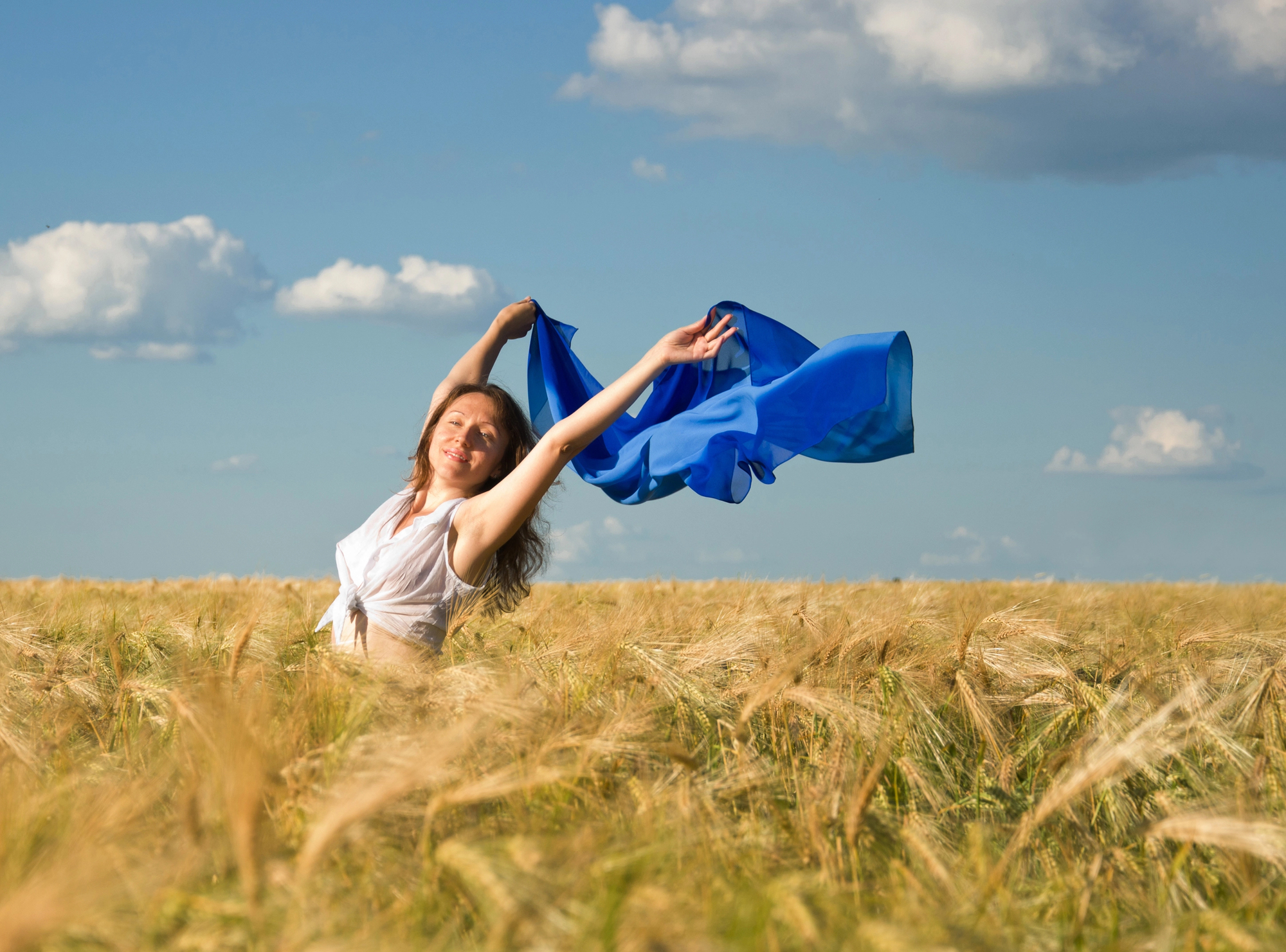 A woman stands in a golden wheat field under a blue sky near Tualatin, holding a flowing blue fabric above her head, smiling joyfully with arms outstretched. Fluffy clouds dot the sky, reflecting the sense of renewal found in Ketamine Therapy.