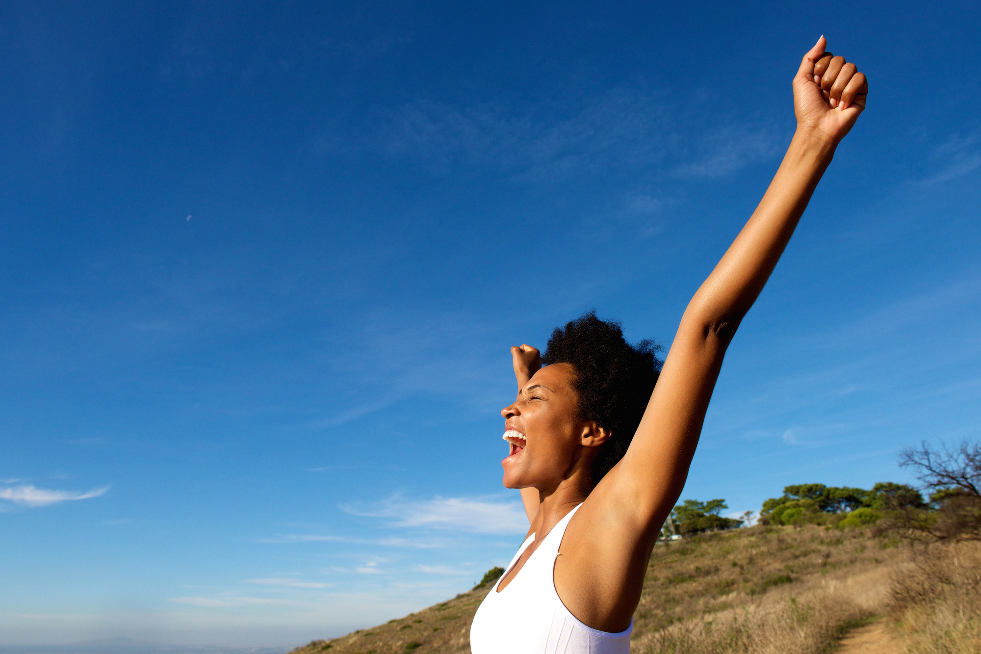 A woman outdoors in King City smiles joyfully with her arms raised in the air under a clear blue sky, standing on a grassy hillside with a few scattered trees in the background.