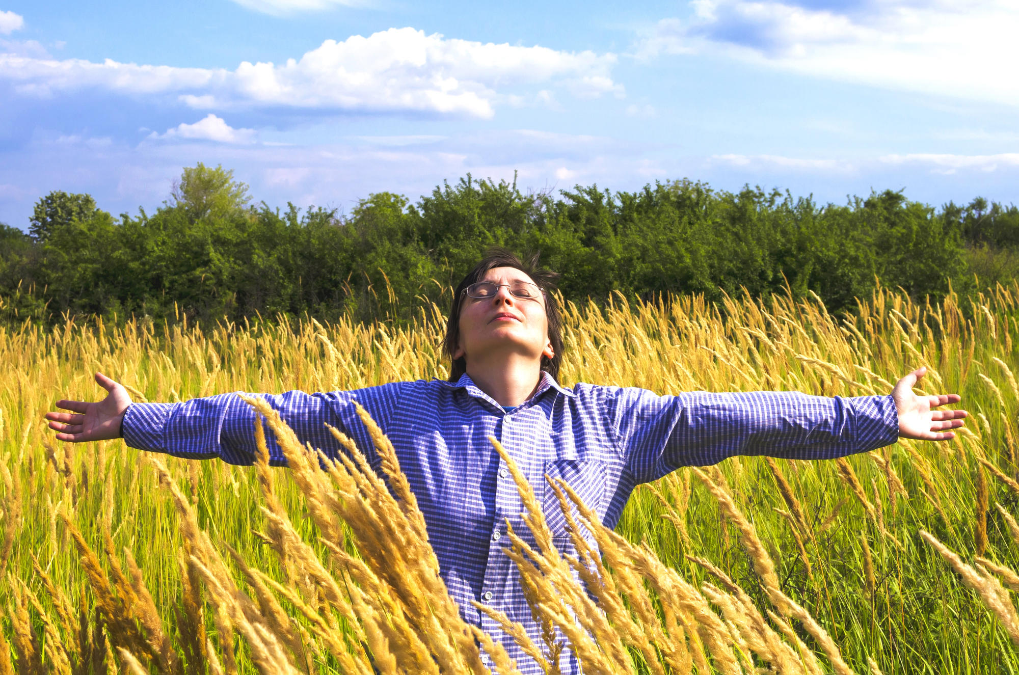 A person in a blue checkered shirt stands in a field of tall yellow grass near Tualatin, arms outstretched, eyes closed, and head tilted upward beneath a partly cloudy sky. Green trees are visible in the background.