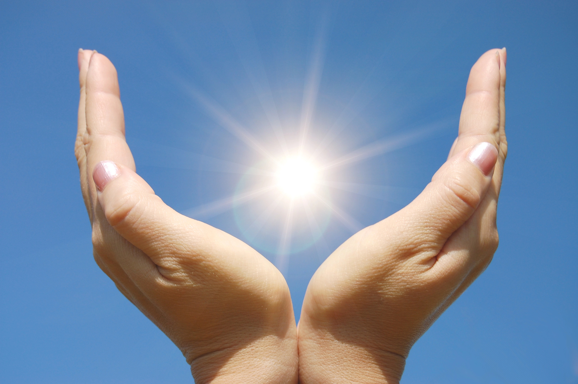 Two hands held close together, cupped upward against a clear blue sky in Vancouver, appear to cradle the bright sun, with sunlight radiating and creating a glowing effect between the palms.