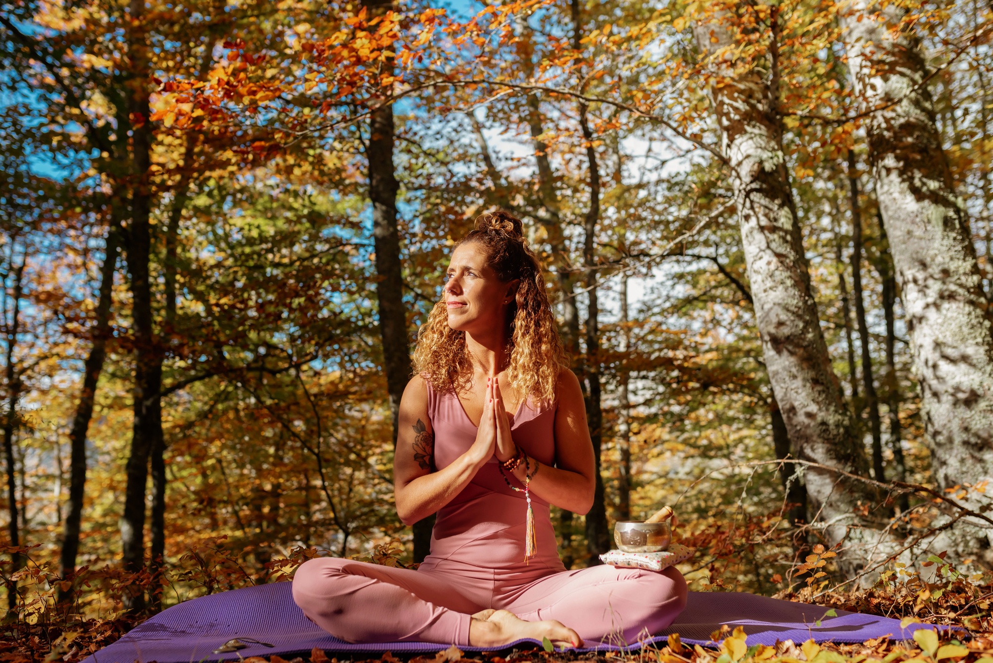 A woman in pink activewear sits cross-legged on a yoga mat in a sunlit King City autumn forest, hands in prayer position, smiling peacefully. Colorful fall leaves surround her, and a singing bowl—often used in ketamine therapy—rests nearby.