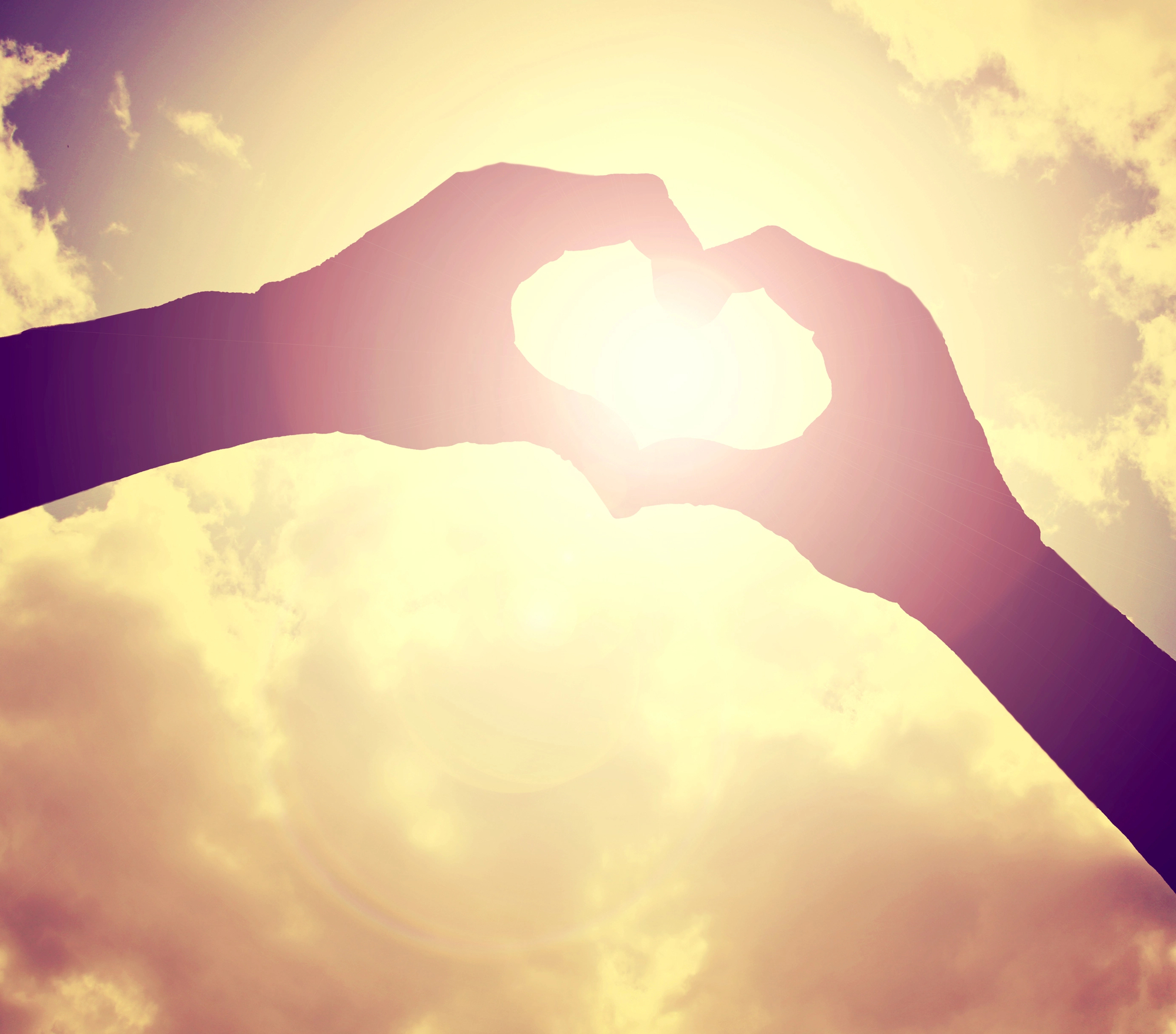 Two hands form a heart shape against a bright, glowing sun with a cloudy sky in the background, creating a silhouette effect—capturing a moment of warmth and hope reminiscent of healing found through Sherwood Ketamine Therapy.