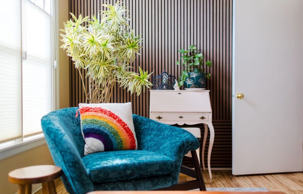 A cozy room with a blue velvet chair, a rainbow pillow, lush potted plants, and wood paneling on the wall offers a serene setting for ketamine therapy, with natural light streaming in from a large window.