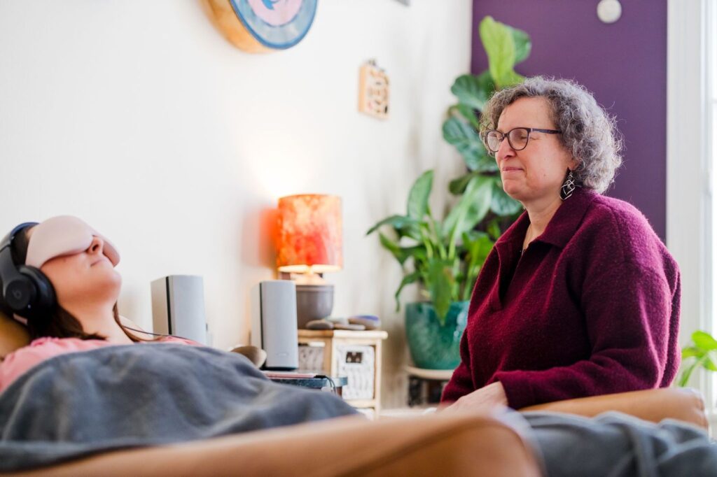 A person lounges under a blanket wearing headphones and an eye mask, experiencing ketamine therapy, while another person with curly hair and glasses sits nearby, observing in a cozy, plant-filled room with soft lighting.