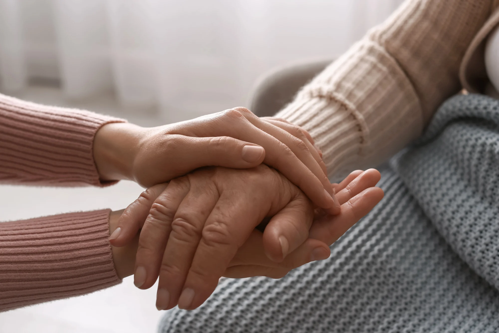 Close-up of a younger person gently holding the hands of an older person, showing care and support. Both wear cozy sweaters, with a blue textured blanket in the background—evoking the compassion found in Ketamine Therapy Tigard.