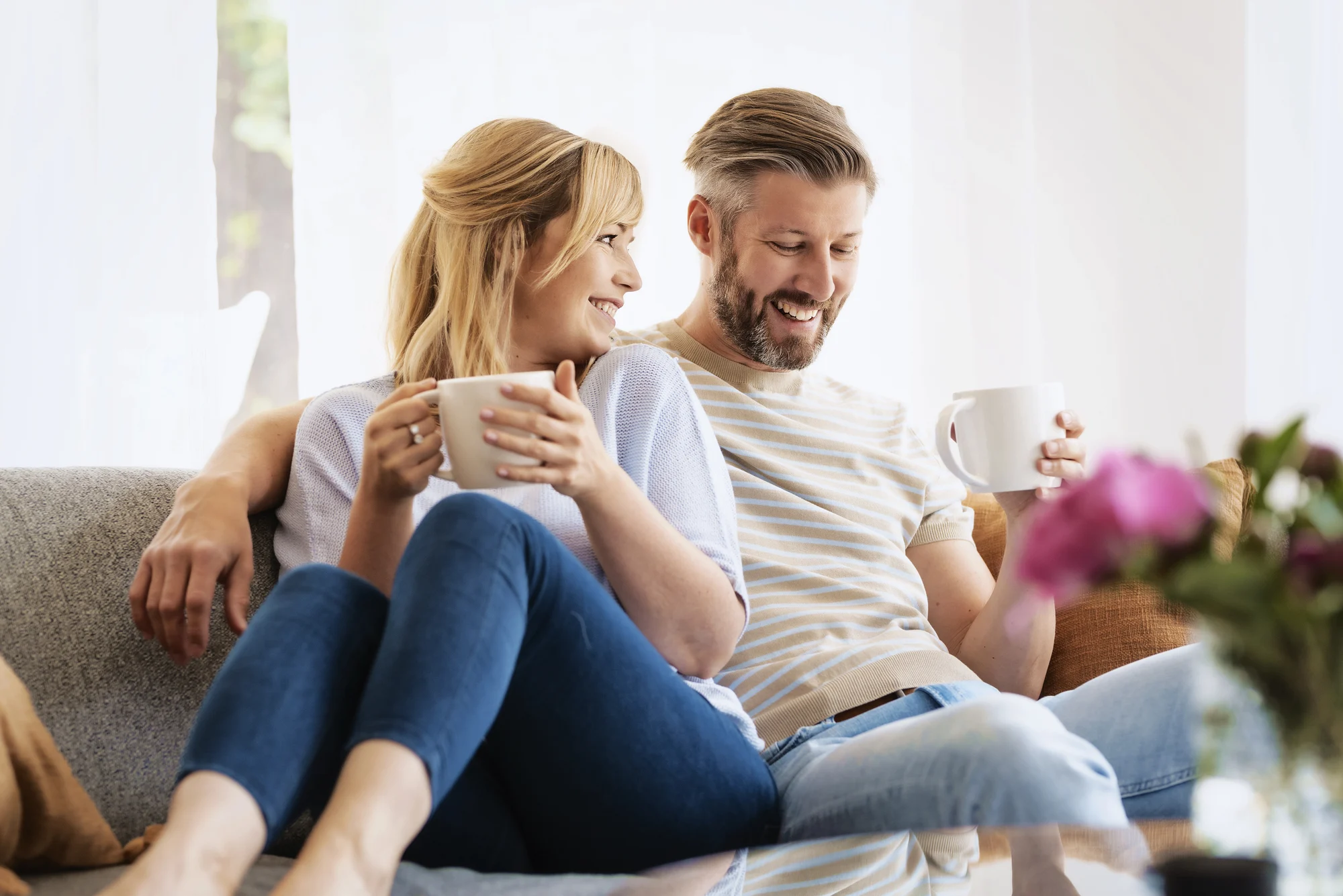 A smiling couple sits closely together on a couch in their Tigard home, each holding a mug. They appear relaxed and happy, enjoying a sunlit living room with a vase of pink flowers—perhaps reflecting the peace found through ketamine therapy.