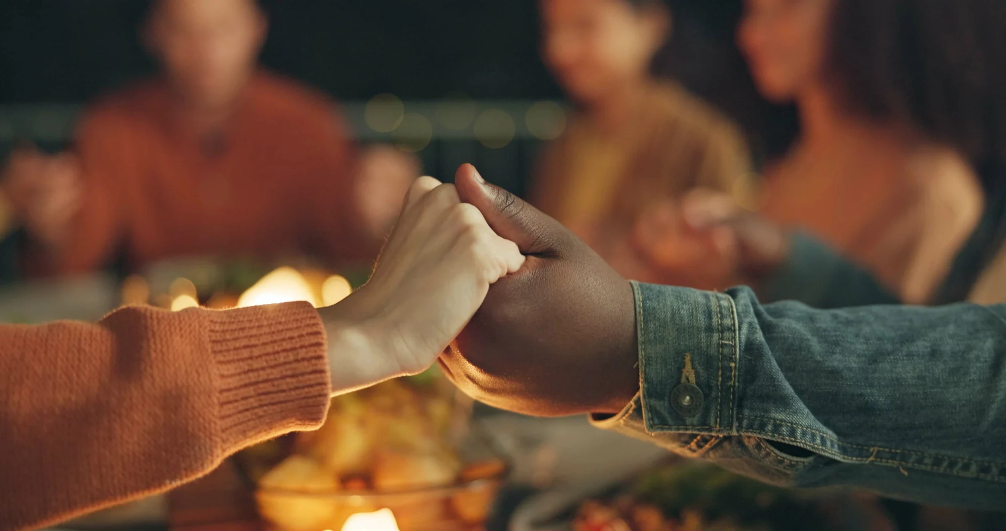 Two people holding hands across a dinner table, with blurred friends in the background sharing a meal in a warm, cozy setting—an inspiring moment of empathy that brings circles of connection closer together.