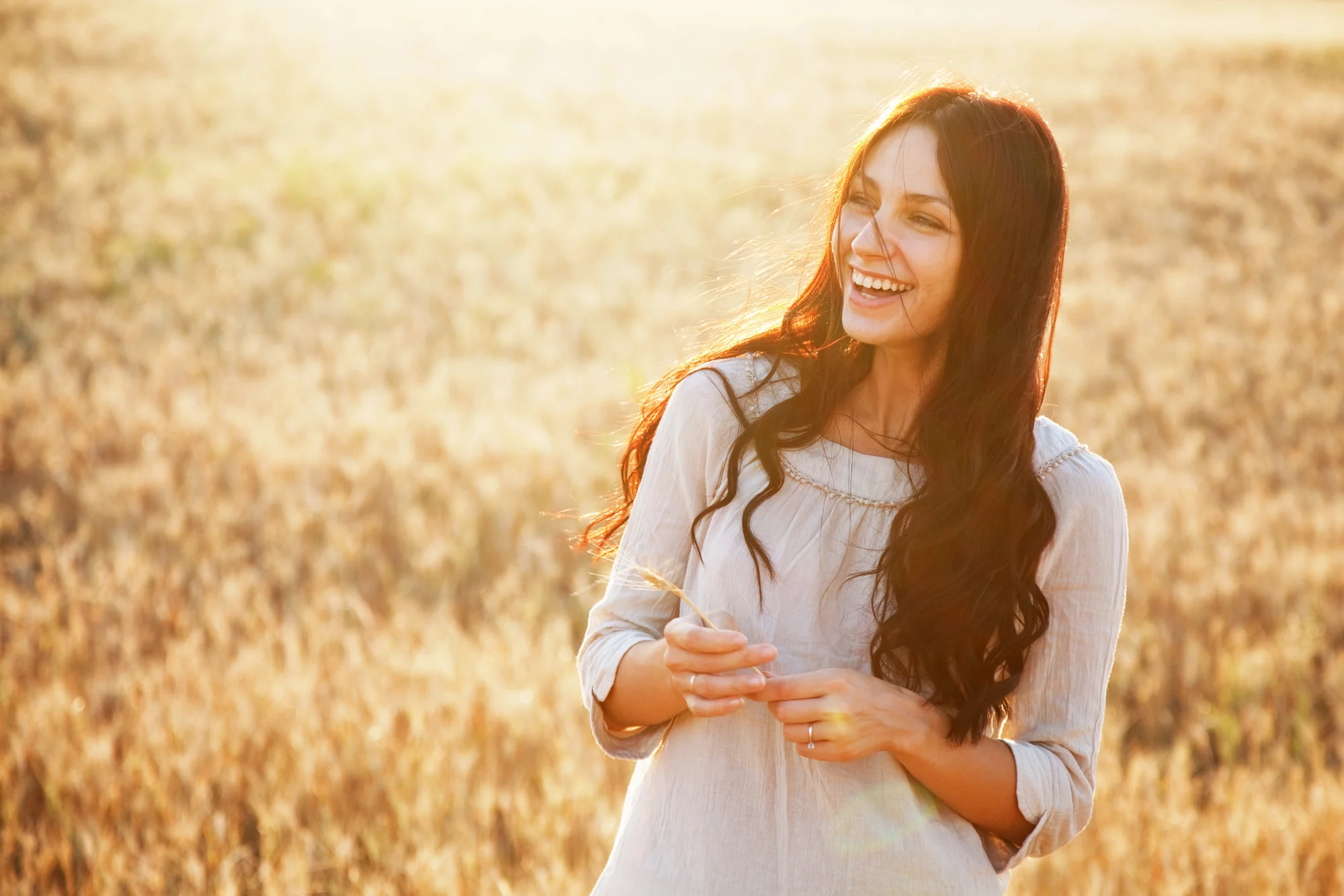 A young woman with long brown hair smiles while standing in a sunlit field of tall grass near Gresham, wearing a light-colored blouse. The background is warmly lit by the sun, evoking the calm often found after Ketamine Therapy.