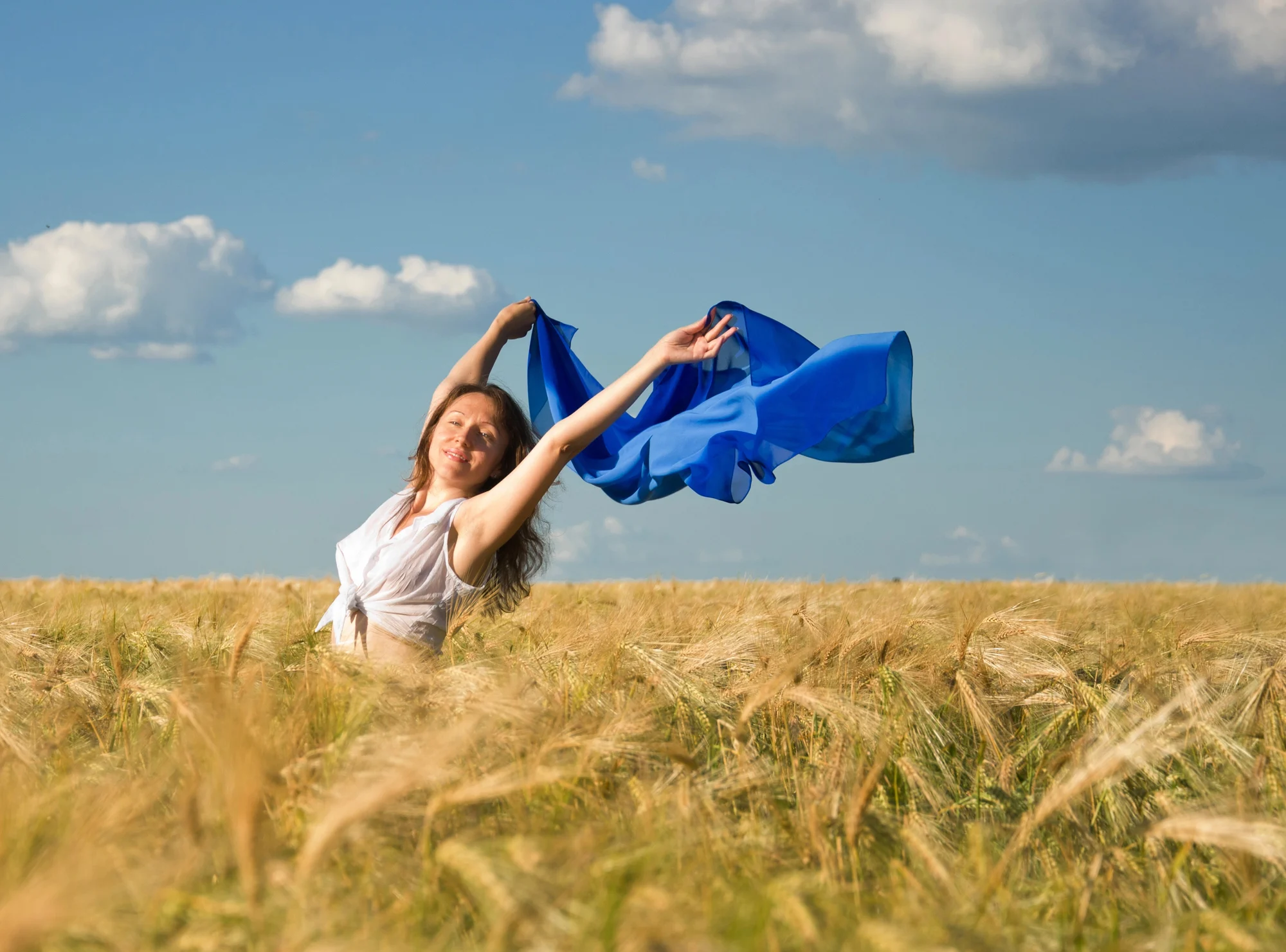 A woman stands in a golden wheat field under a blue sky near Tualatin, holding a flowing blue fabric above her head, smiling joyfully with arms outstretched. Fluffy clouds dot the sky, reflecting the sense of renewal found in Ketamine Therapy.