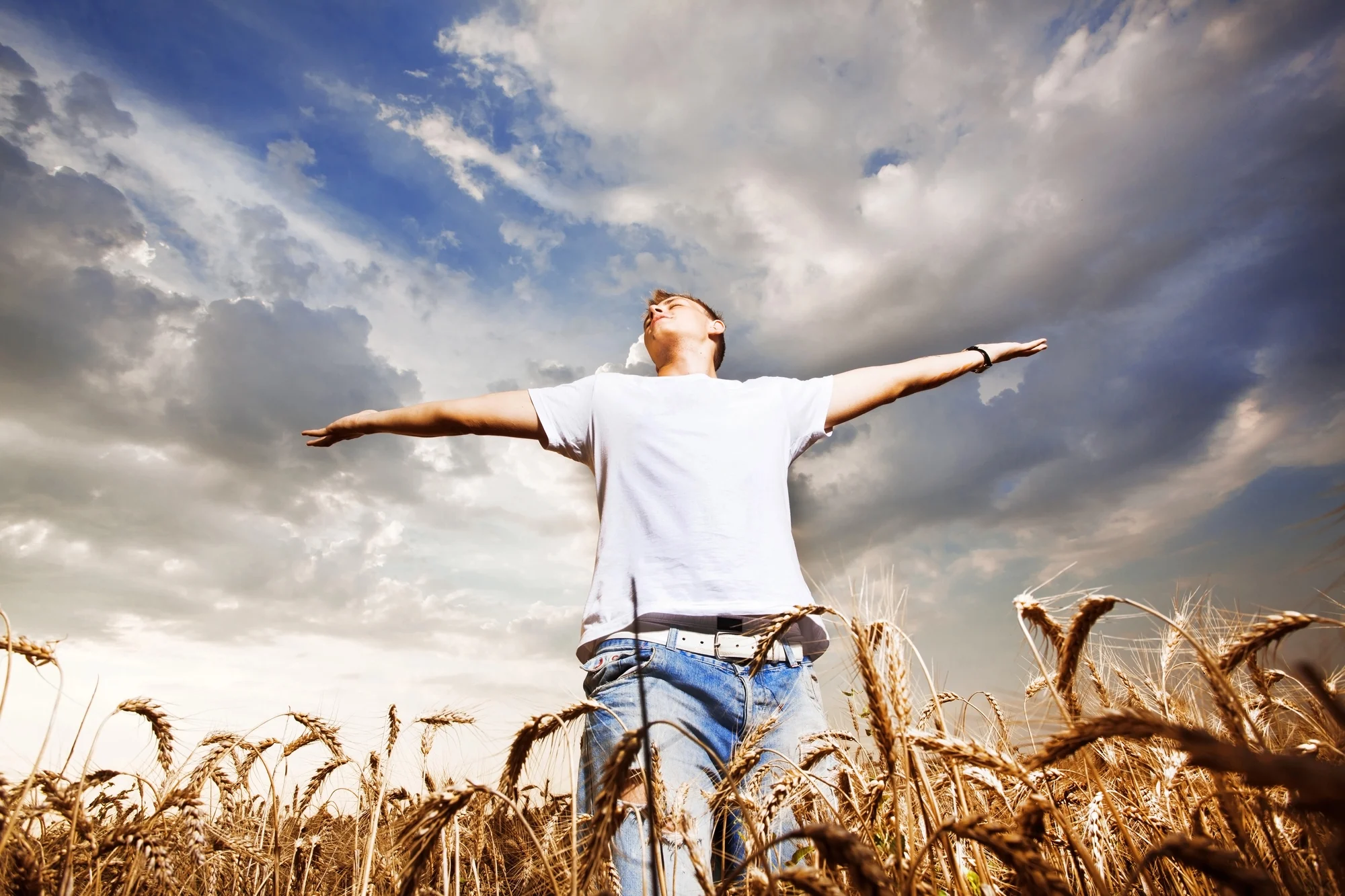 A person in a white t-shirt and jeans stands in a wheat field with arms outstretched, looking up at a dramatic, cloudy sky, exuding joyful, aloha-inspired freedom—like embracing life after transformative ketamine therapy.