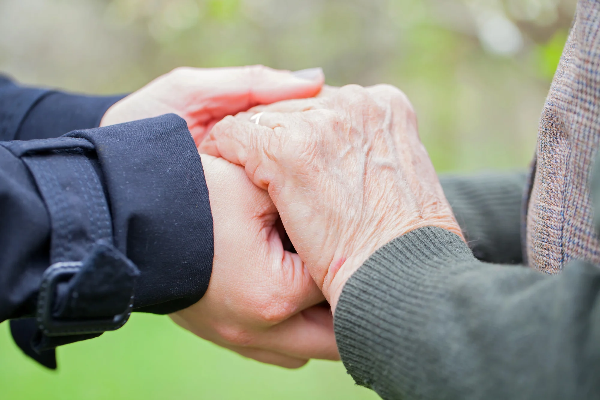 A younger person gently holds the hands of an older person outdoors in Oregon City, showing care and support. The older person is wearing a green sweater, the younger a dark coat, with blurred greenery in the background.