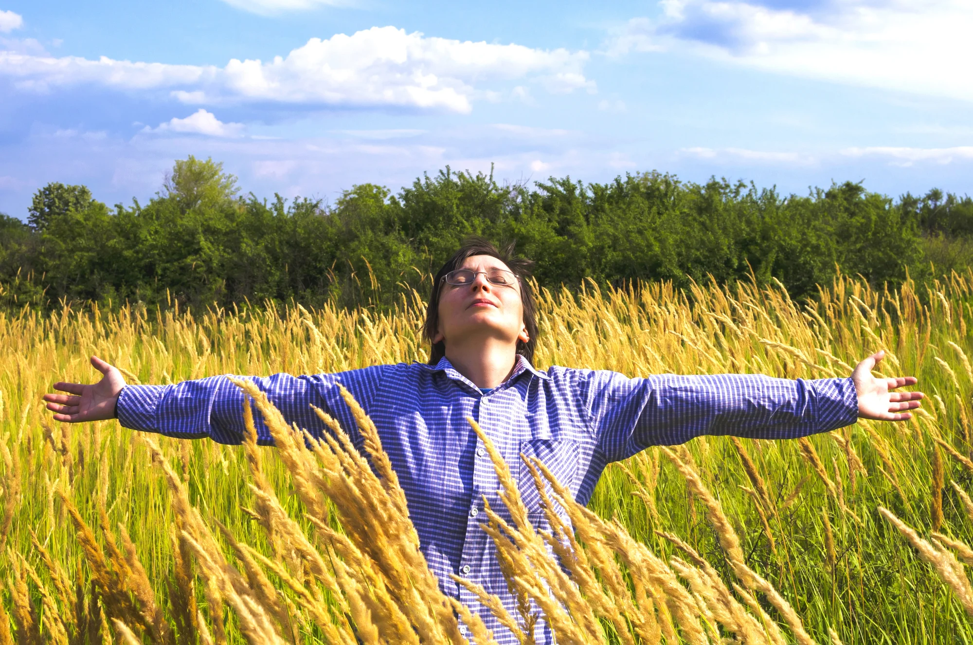 A person in a blue checkered shirt stands in a field of tall yellow grass near Tualatin, arms outstretched, eyes closed, and head tilted upward beneath a partly cloudy sky. Green trees are visible in the background.