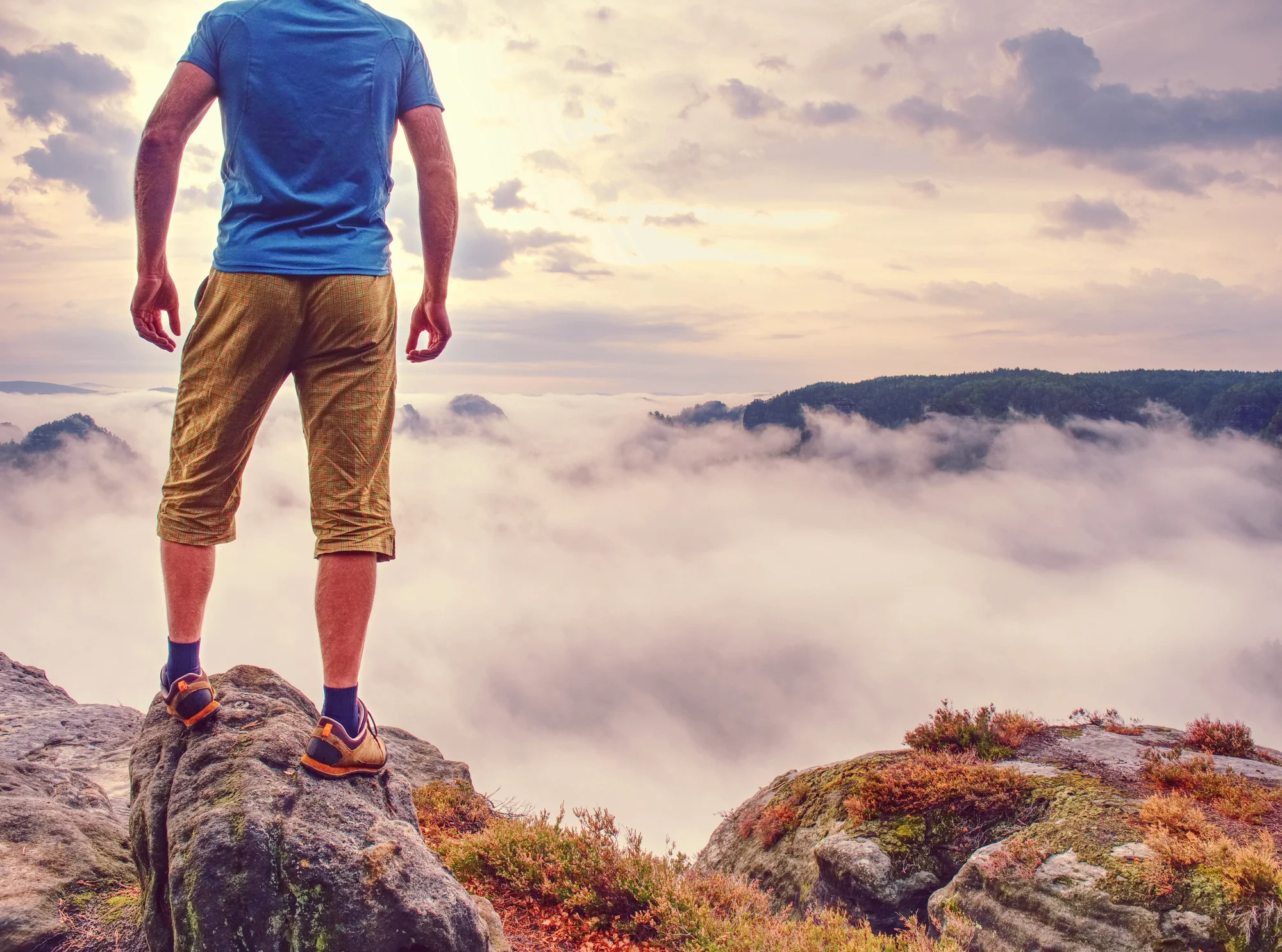 A person stands on a rocky cliff in Washington, overlooking a scenic landscape filled with clouds and mist, with distant hills and a colorful sky at sunrise or sunset.
