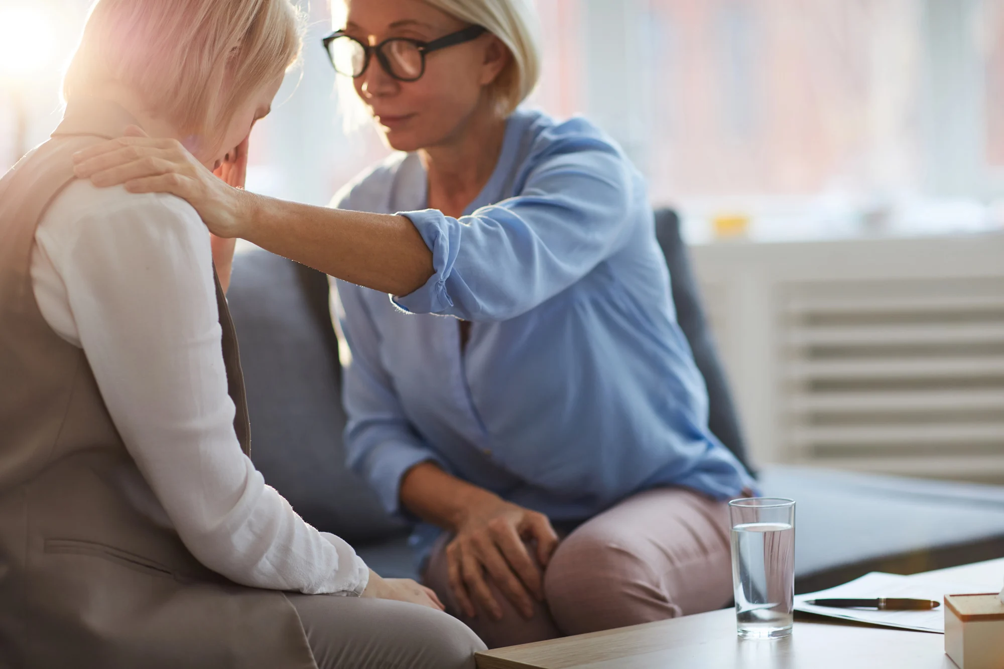 A woman comforts another woman by placing a hand on her shoulder as they sit together on a couch in Vancouver, with soft natural light coming through a window. A glass of water and a notebook, perhaps about Ketamine Therapy, rest on the table nearby.