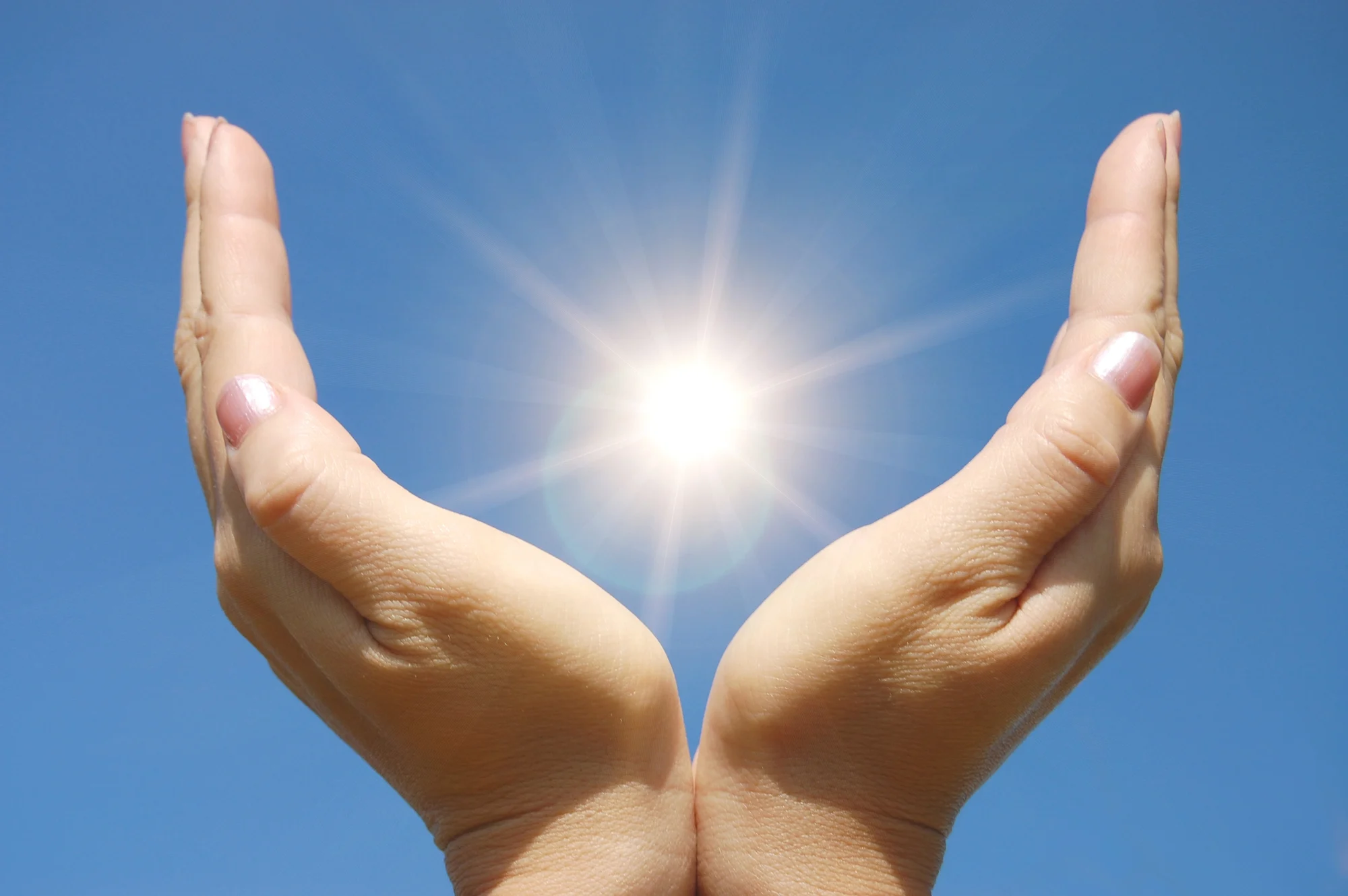 Two hands held close together, cupped upward against a clear blue sky in Vancouver, appear to cradle the bright sun, with sunlight radiating and creating a glowing effect between the palms.
