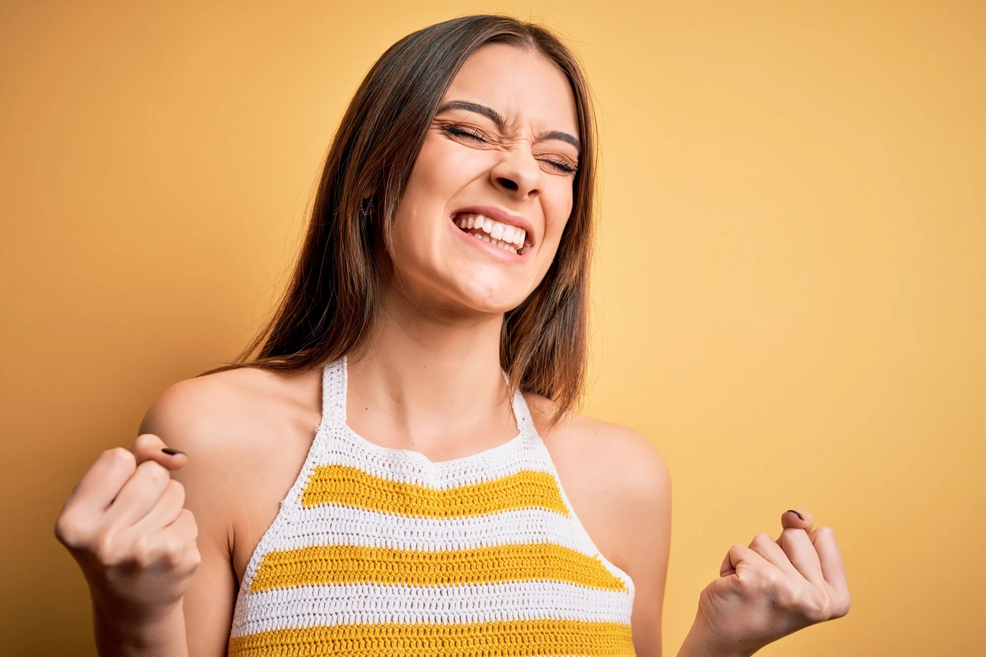 A young woman with long brown hair smiles joyfully with her eyes closed, fists clenched in excitement, wearing a striped yellow and white top against a solid yellow background, radiating pure Aloha spirit.