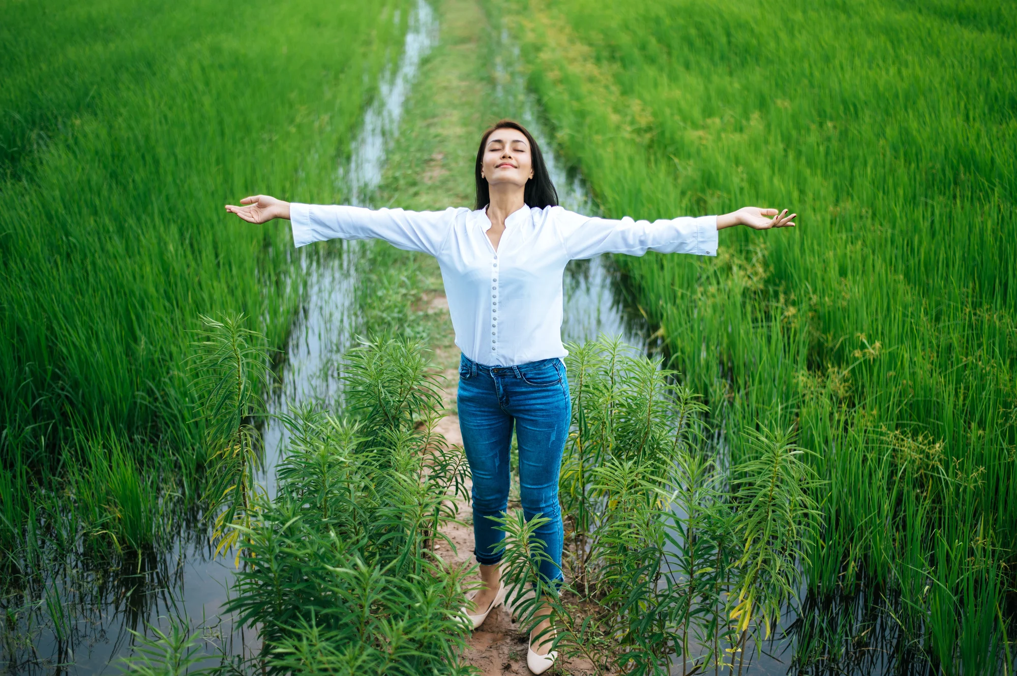 A woman in a white shirt and jeans stands on a narrow path between green fields near Hillsboro, arms outstretched and eyes closed, appearing relaxed and content, as if embracing the calm after ketamine therapy in nature.