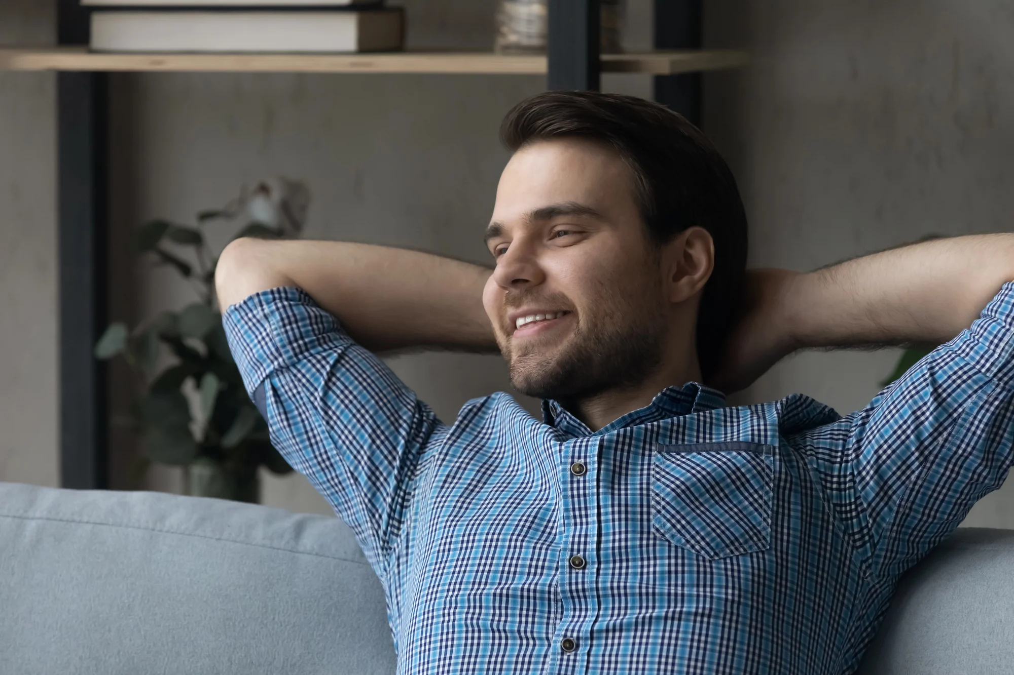 A man in a blue checkered shirt is sitting on a couch, leaning back with his hands behind his head, smiling and looking relaxed—an image that captures the calm often sought through ketamine therapy. Shelves and a plant are visible in the background.