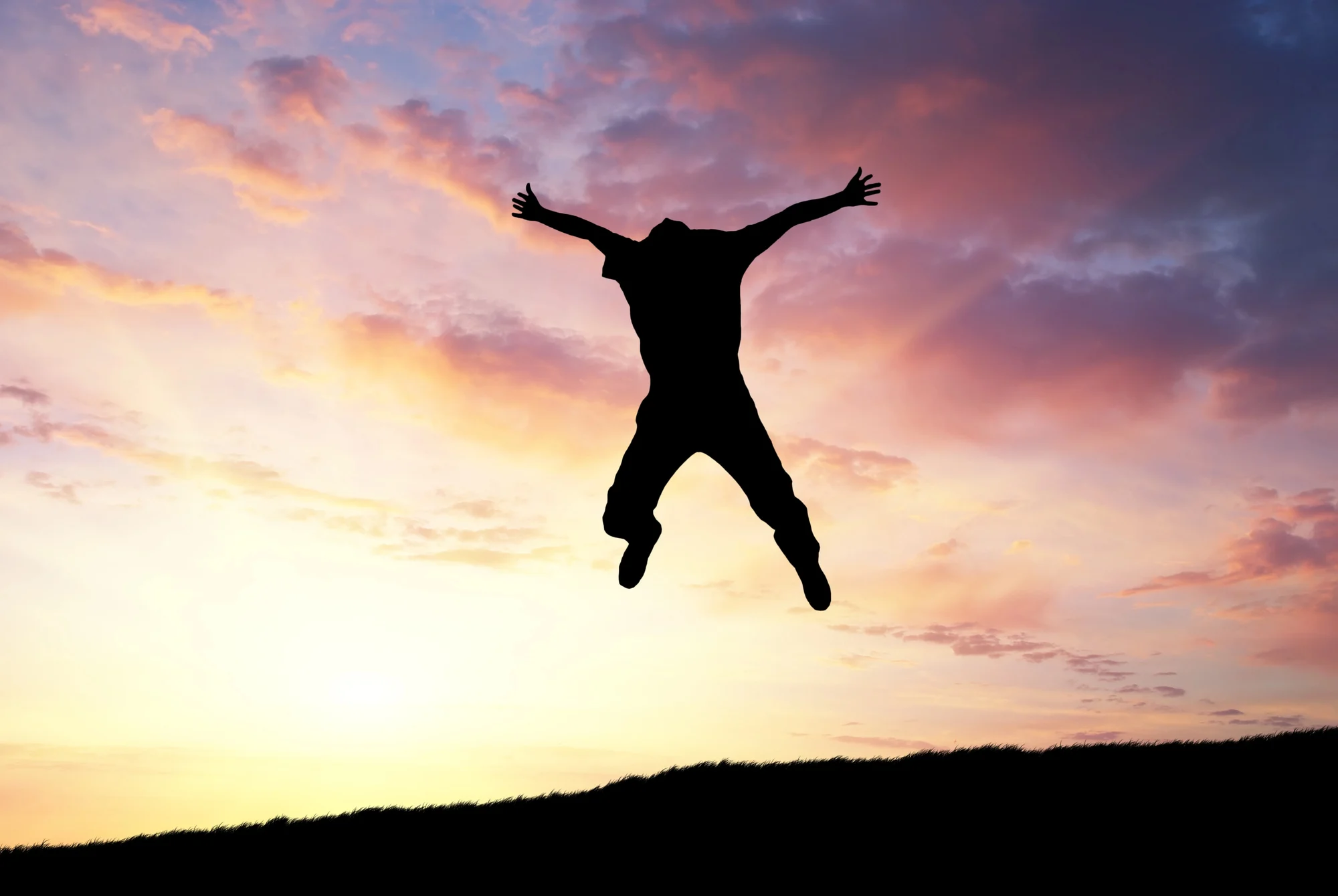 A person is silhouetted against a colorful sunset sky, jumping with arms and legs outstretched on a Happy Valley hilltop.