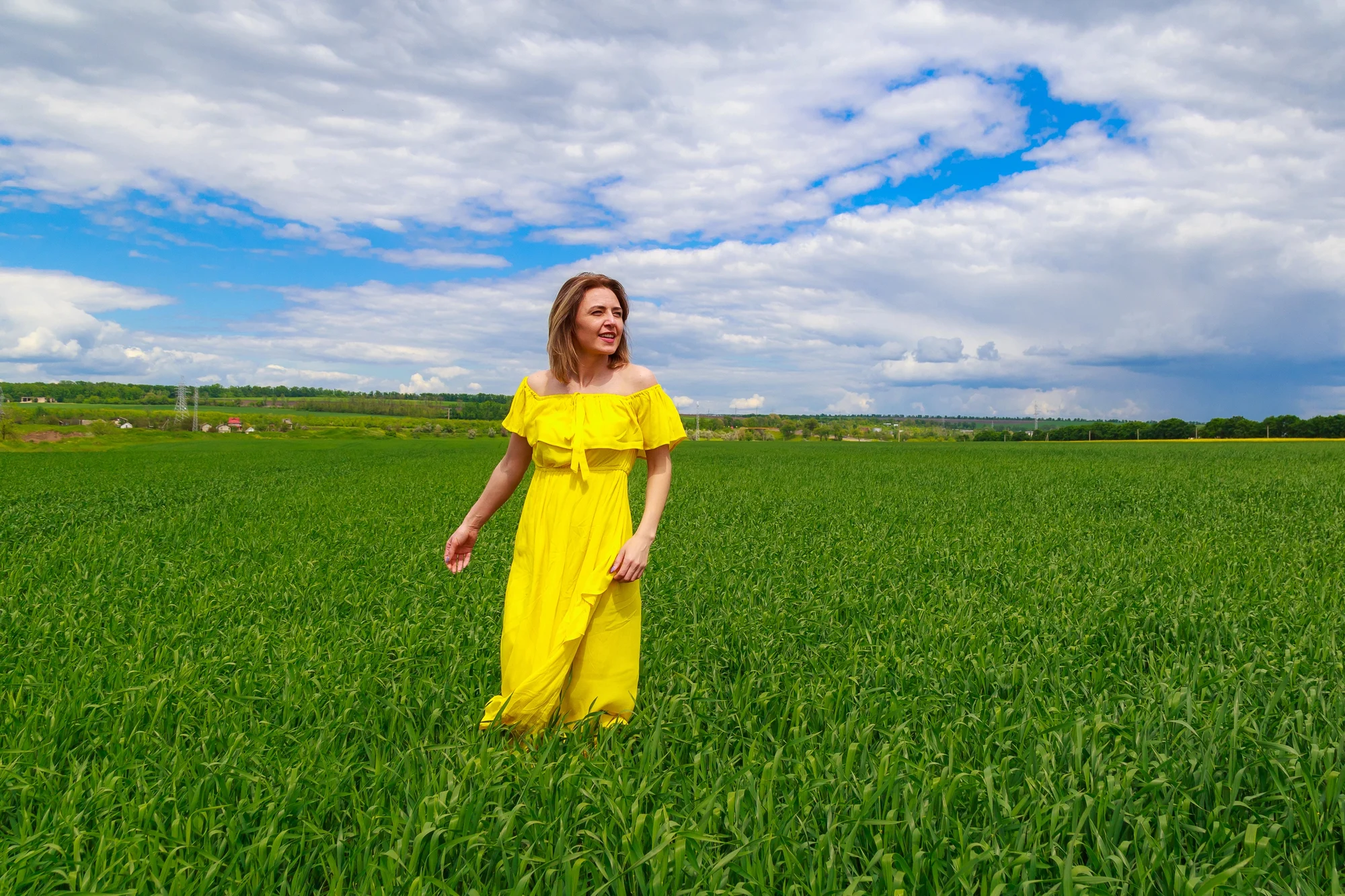 A woman in a bright yellow dress stands in a lush green field near Oregon City under a partly cloudy blue sky, smiling and looking to the side.