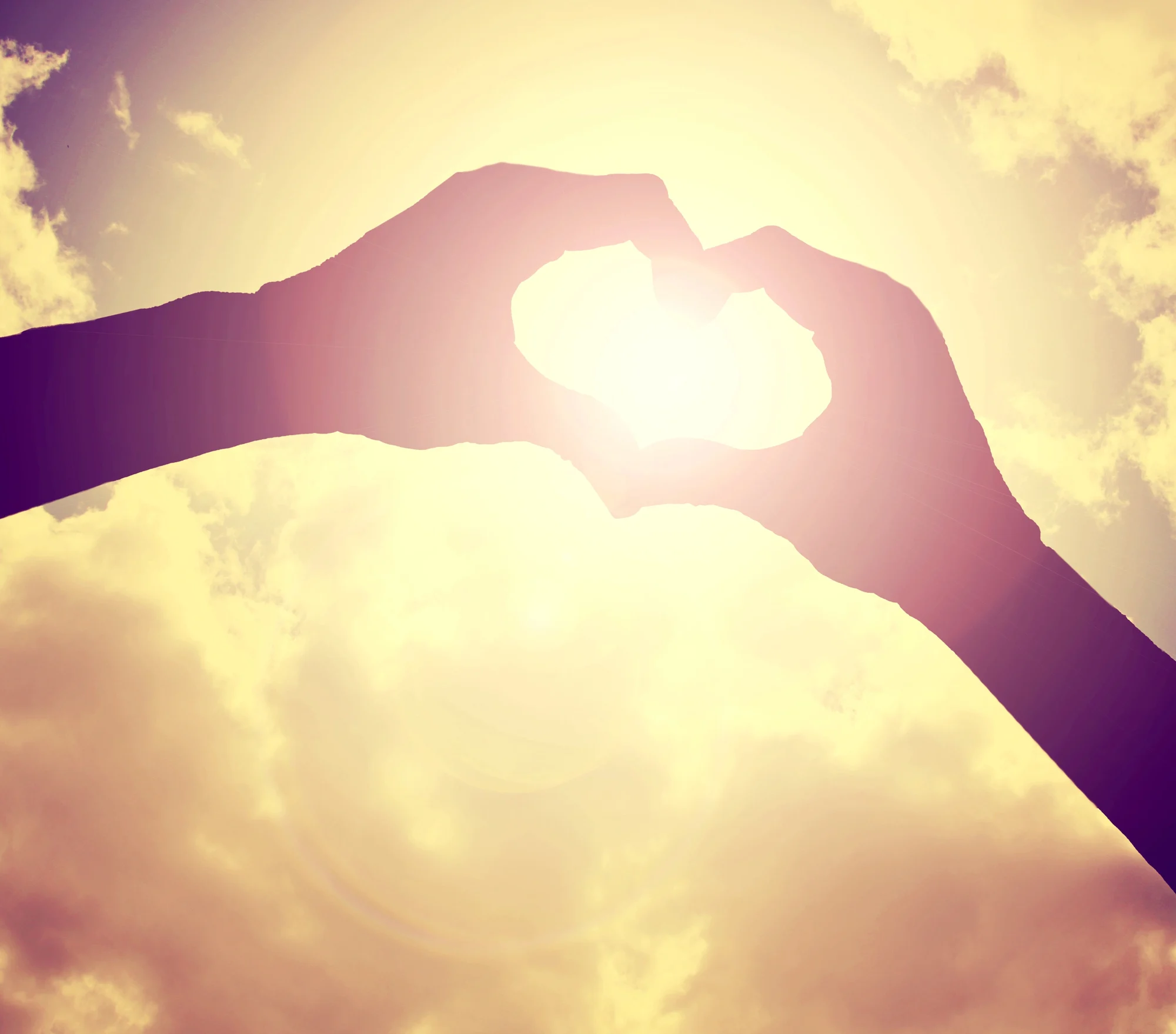 Two hands form a heart shape against a bright, glowing sun with a cloudy sky in the background, creating a silhouette effect—capturing a moment of warmth and hope reminiscent of healing found through Sherwood Ketamine Therapy.