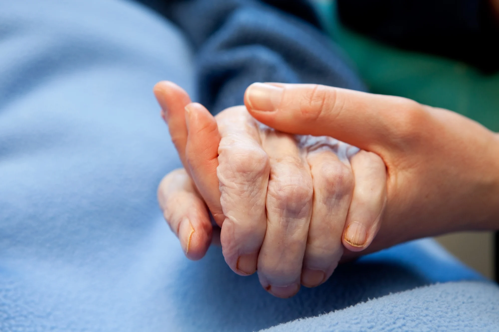 A younger person gently holds the hand of an older person, both resting on a light blue blanket in Happy Valley, conveying care, support, and compassion.