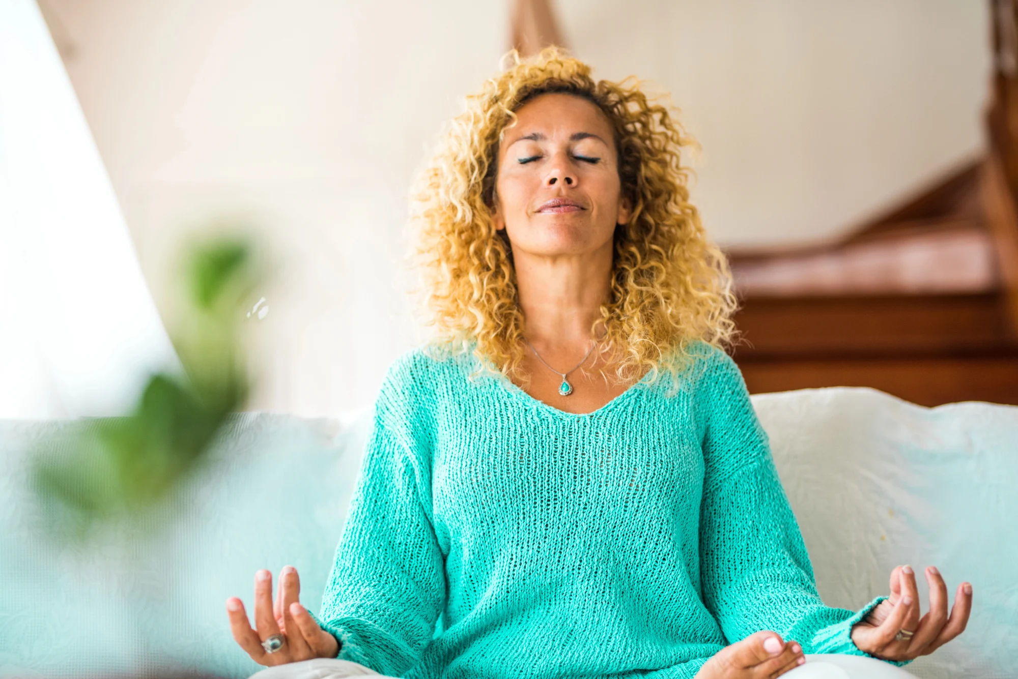 A woman with curly blonde hair sits cross-legged on a couch in Gresham, eyes closed, wearing a turquoise sweater, meditating with relaxed hands in her lap in a brightly lit room after Ketamine Therapy.
