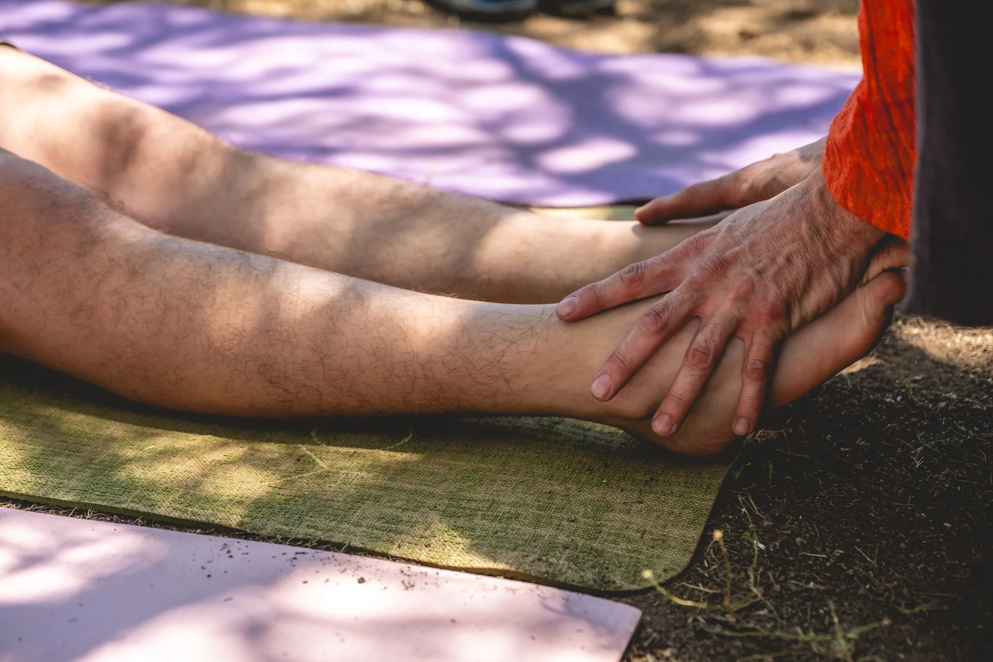 Close-up of a person’s hands gently massaging another person's lower legs and ankles outdoors on a mat at Cedar Hills, with dappled sunlight and shadows visible on the ground—evoking the serenity often sought in ketamine therapy sessions.