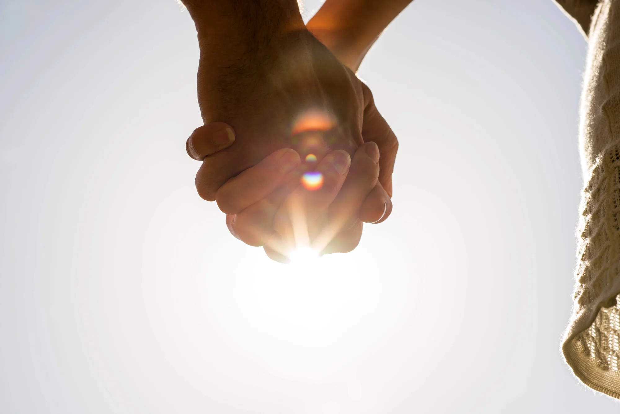 Two people holding hands with sunlight shining between their fingers, creating a lens flare effect and bright, warm atmosphere. Captured in Tualatin, the image emphasizes unity and connection—much like shared journeys in ketamine therapy.