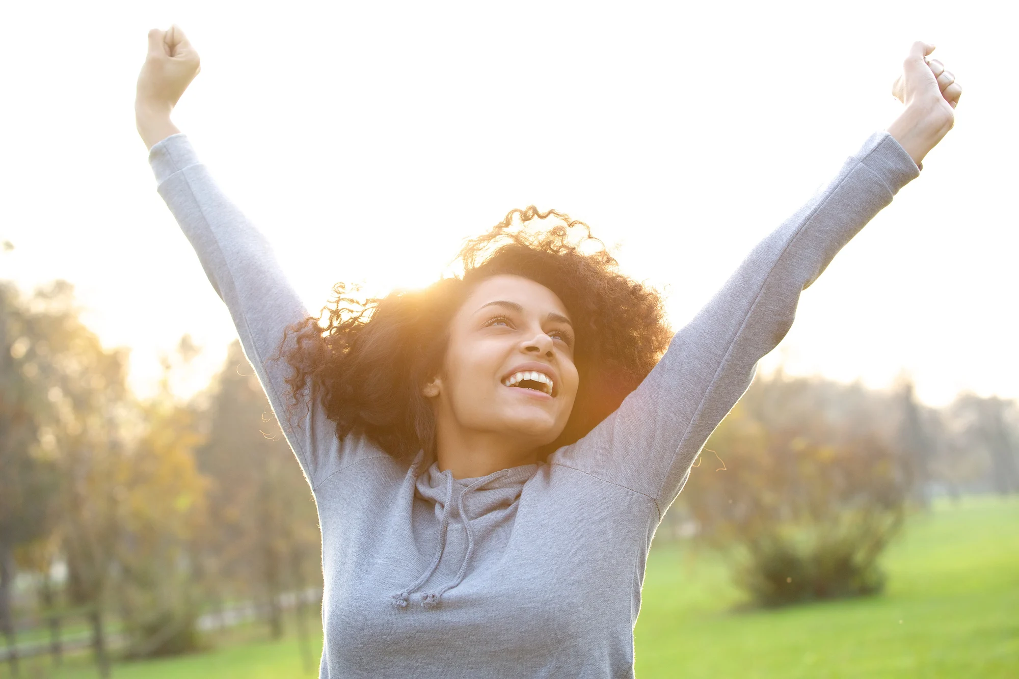 A young woman outdoors in Sherwood park, smiling joyfully with both arms raised in the air. She is wearing a gray hoodie and is backlit by bright sunlight, with trees and green grass in the background, embodying hope like Ketamine Therapy.