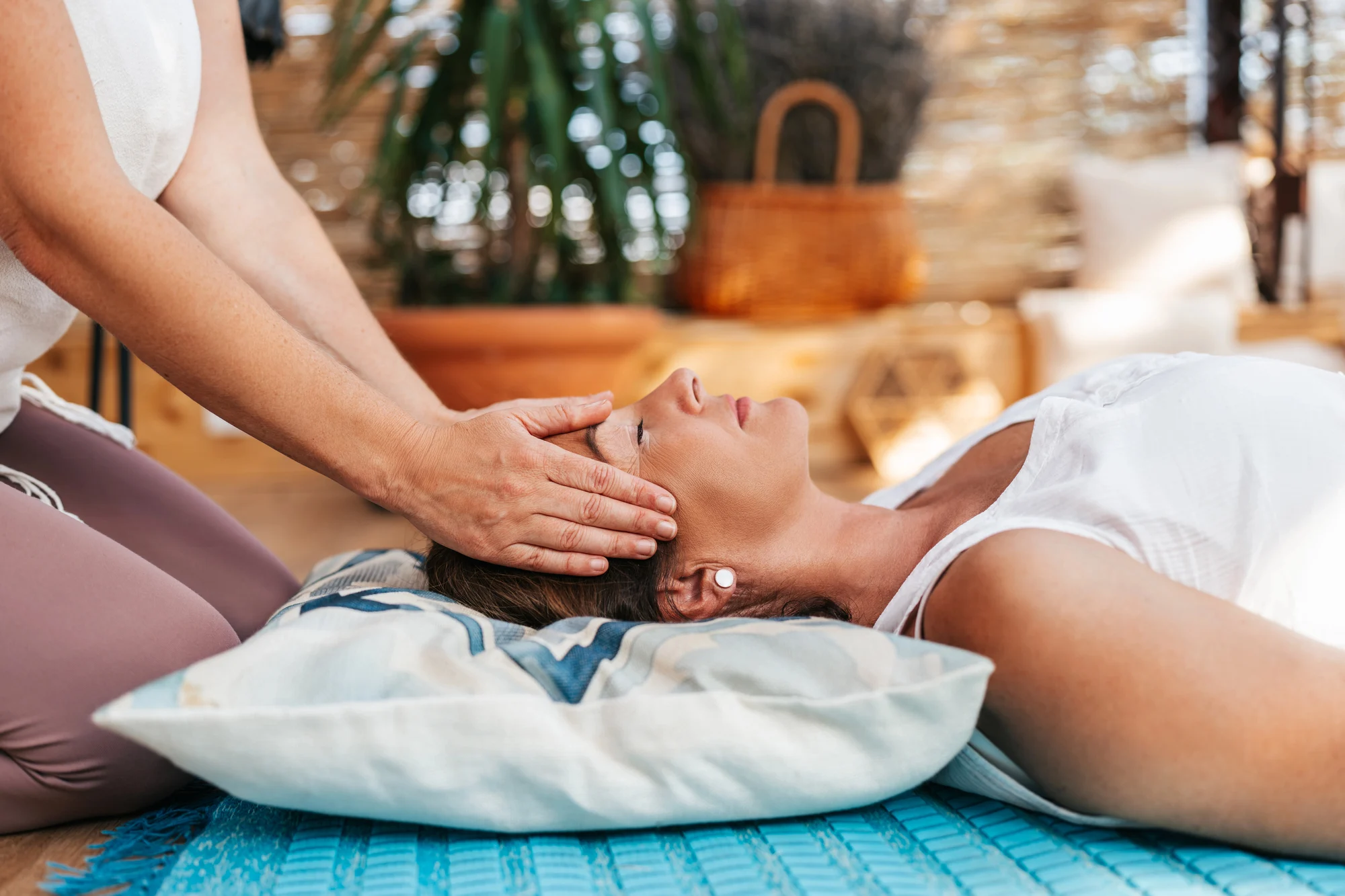A woman lies on a pillow with her eyes closed while another person gently places their hands on her head, suggesting a relaxing Ketamine Therapy session in a peaceful, indoor setting infused with an Aloha spirit.