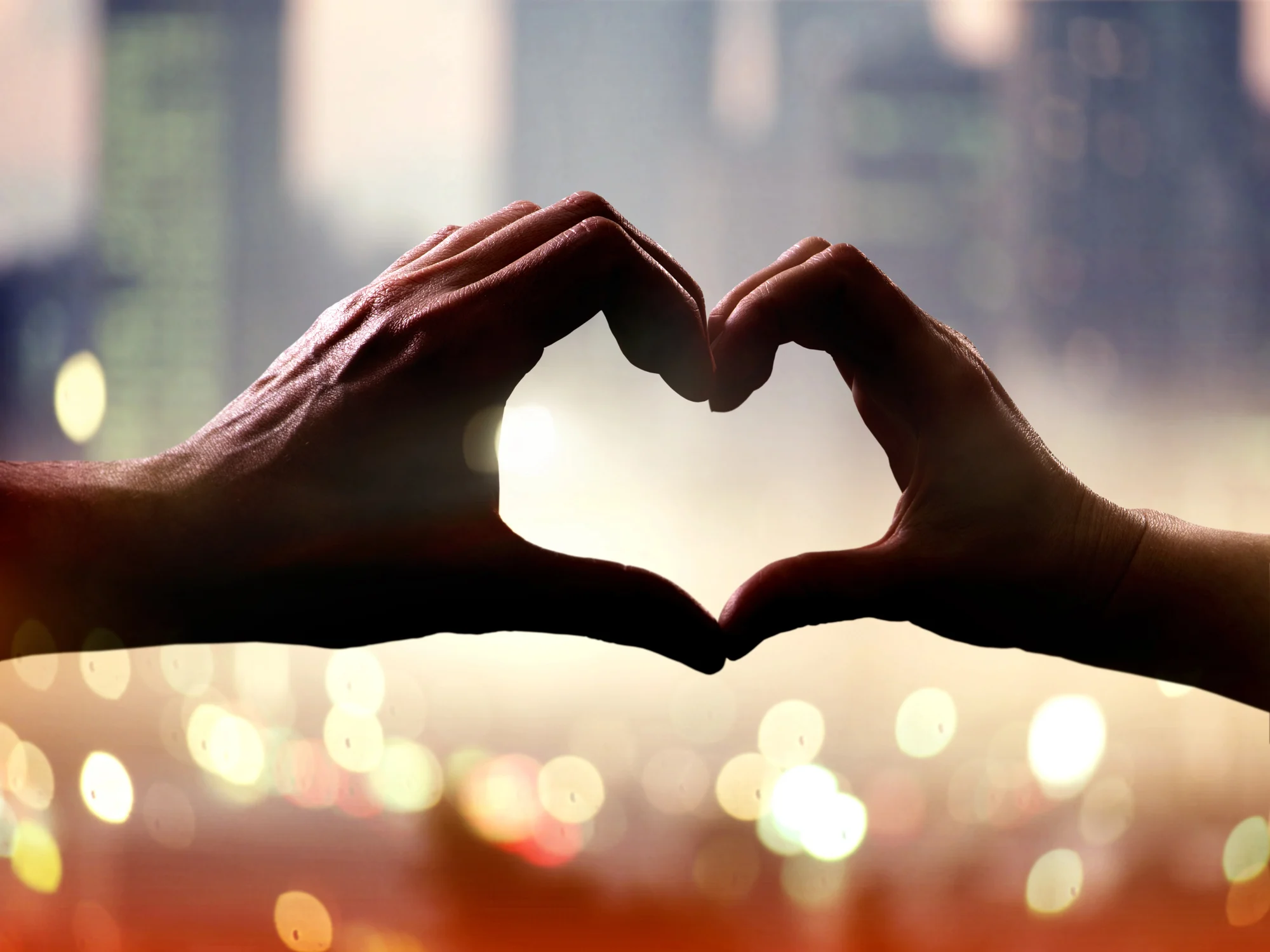 Two hands forming the shape of a heart against a blurred city skyline with warm, glowing lights in the background, capturing the uplifting spirit often found in Happy Valley or after transformative ketamine therapy.